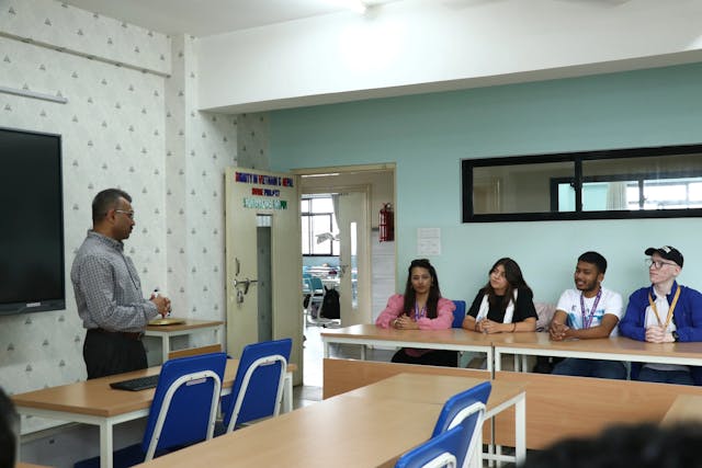A picture where Fr. Jiju is giving a welcome speech and addressing the dedicated team. On the right, Sanjiya, Victoria, Abhishek and Joon can be observed sitting.