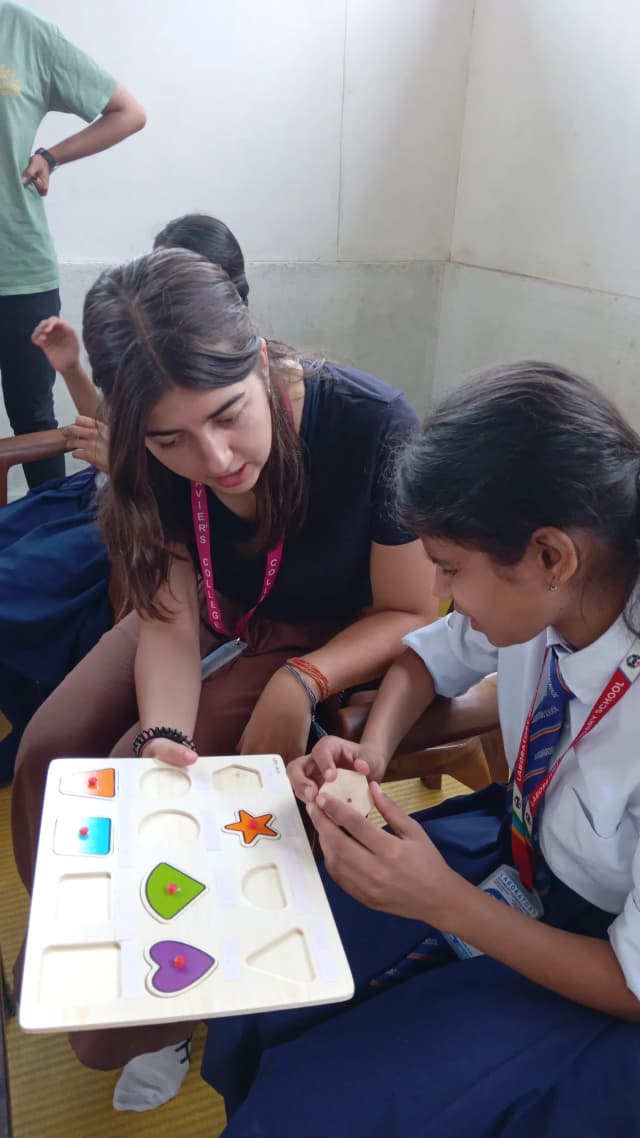 A girl child is perceiving one of the 2D wooden geometrical shapes and the international intern Victoria is holding the wooden board.