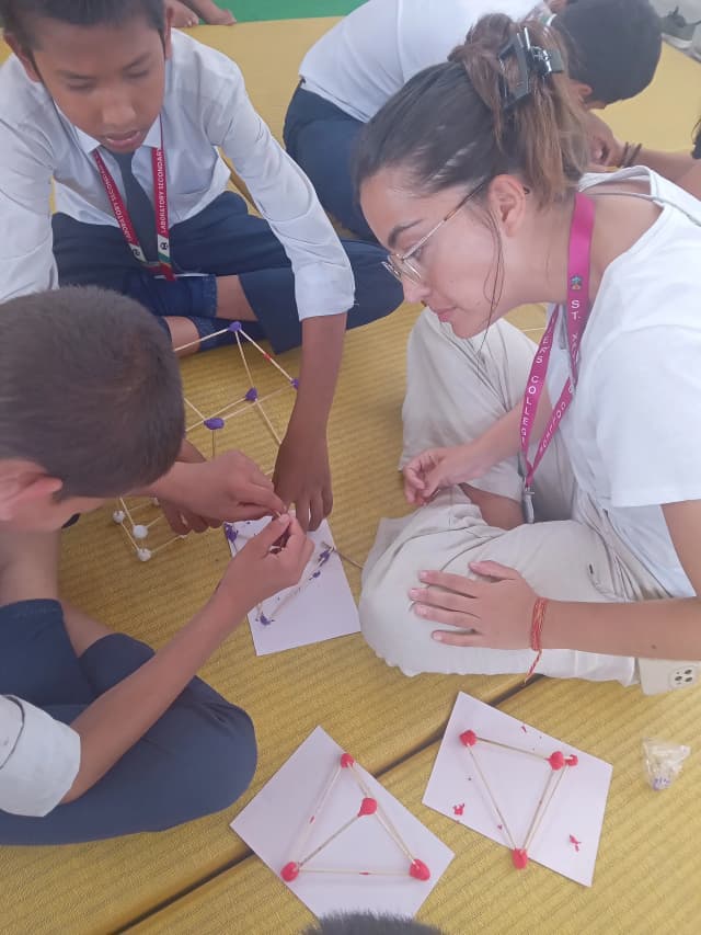 2 of the children are making 3D geometrical shapes using clay and sticks and International Intern Valeria is observing them.