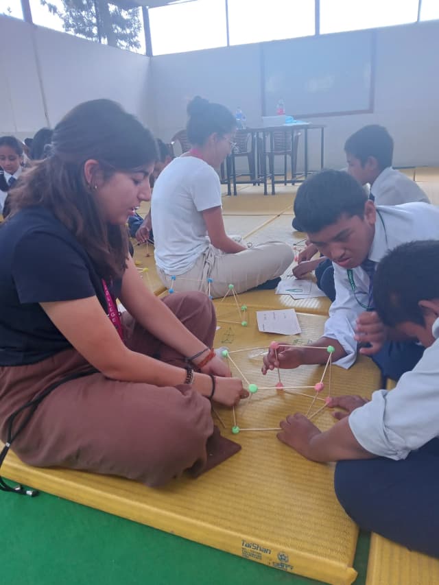 2 of the children are making 3D geometrical shapes using clay and sticks and International Intern Victoria is observing them.