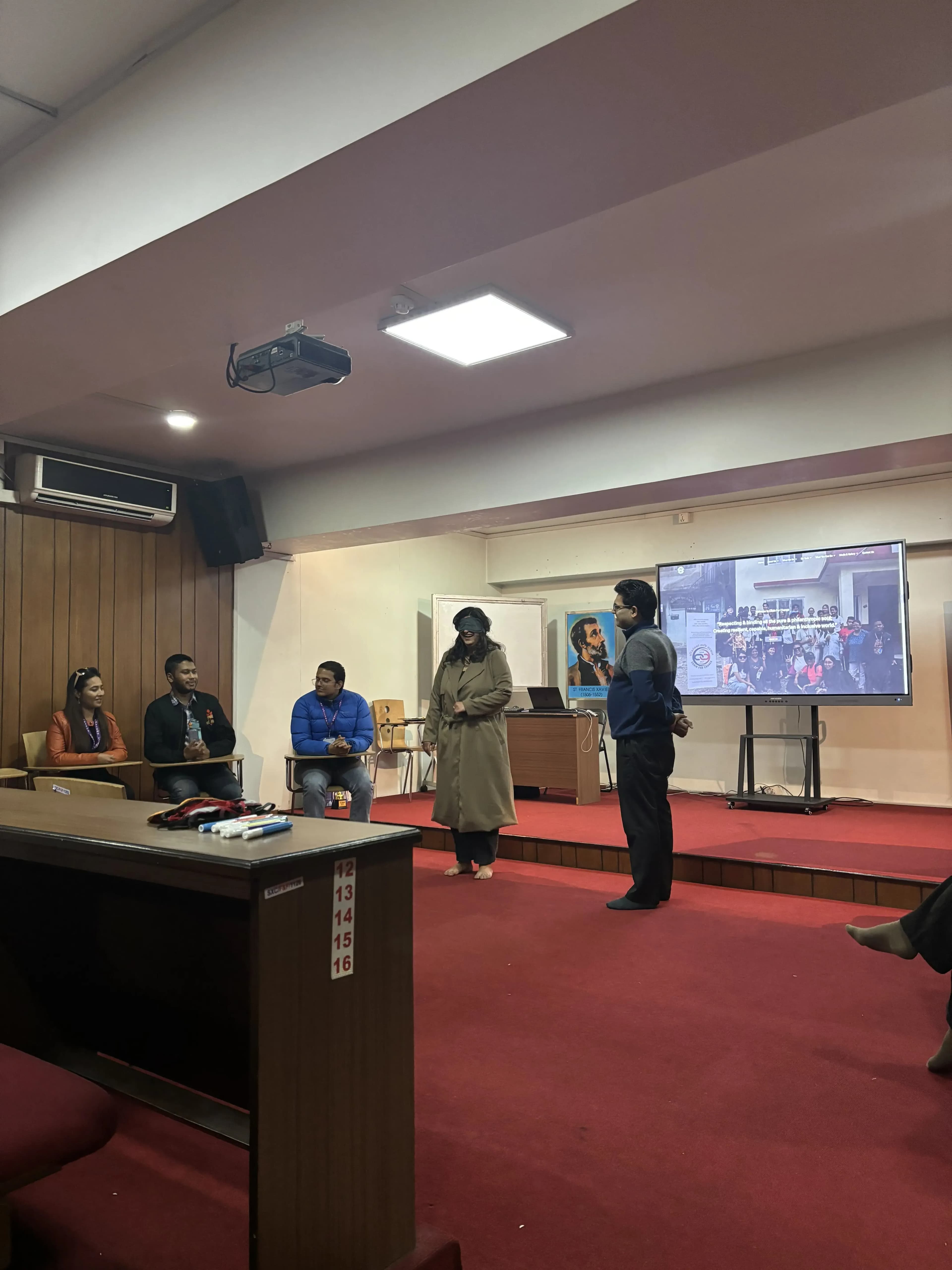 A female participant is being blindfolded and a male participant is standing beside her about to be ready for the instruction of the game. In the background Founders of CFC and Project members are observed and on the screen the website of CFC is being displayed.