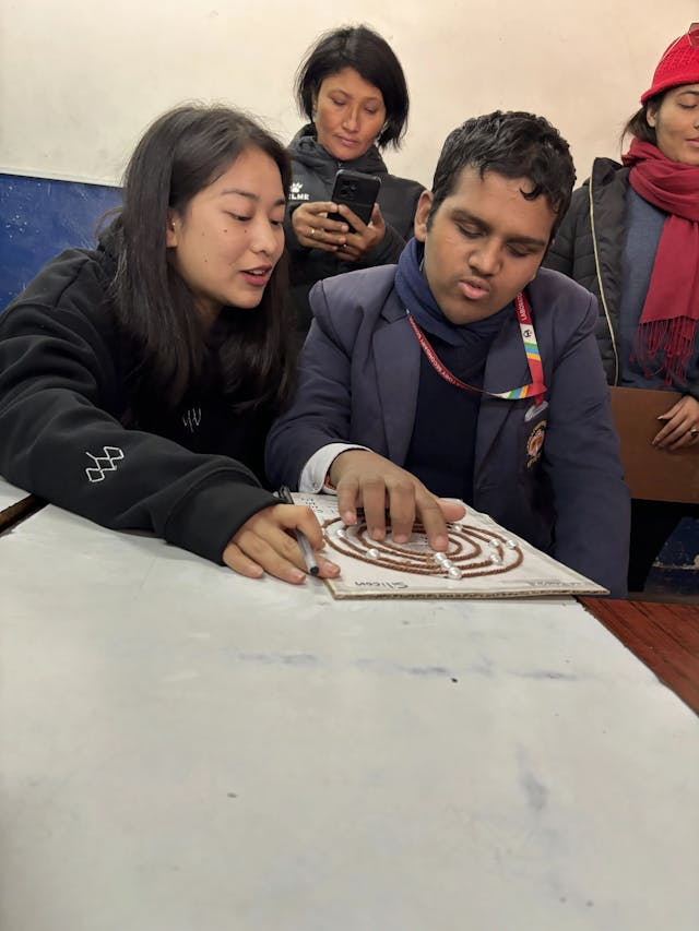 A male participant is perceiving the 3D model of atomic structure and Sweta Shakya - project member is explaining him. In the background bina Shrestha is observed using her phone.