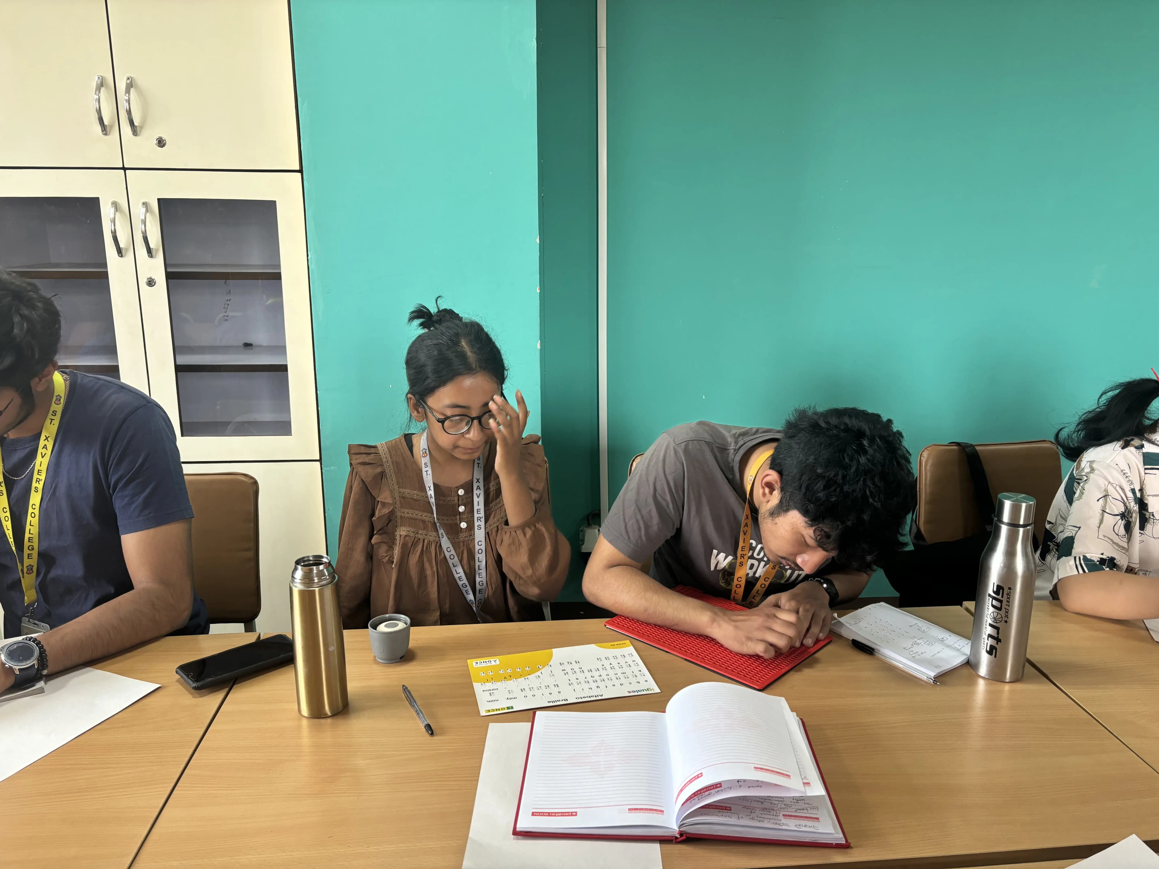 On right, Project member Aakash Shrestha is writing braille using slate and stylus and on left Project Member Garima Shakya is observed.