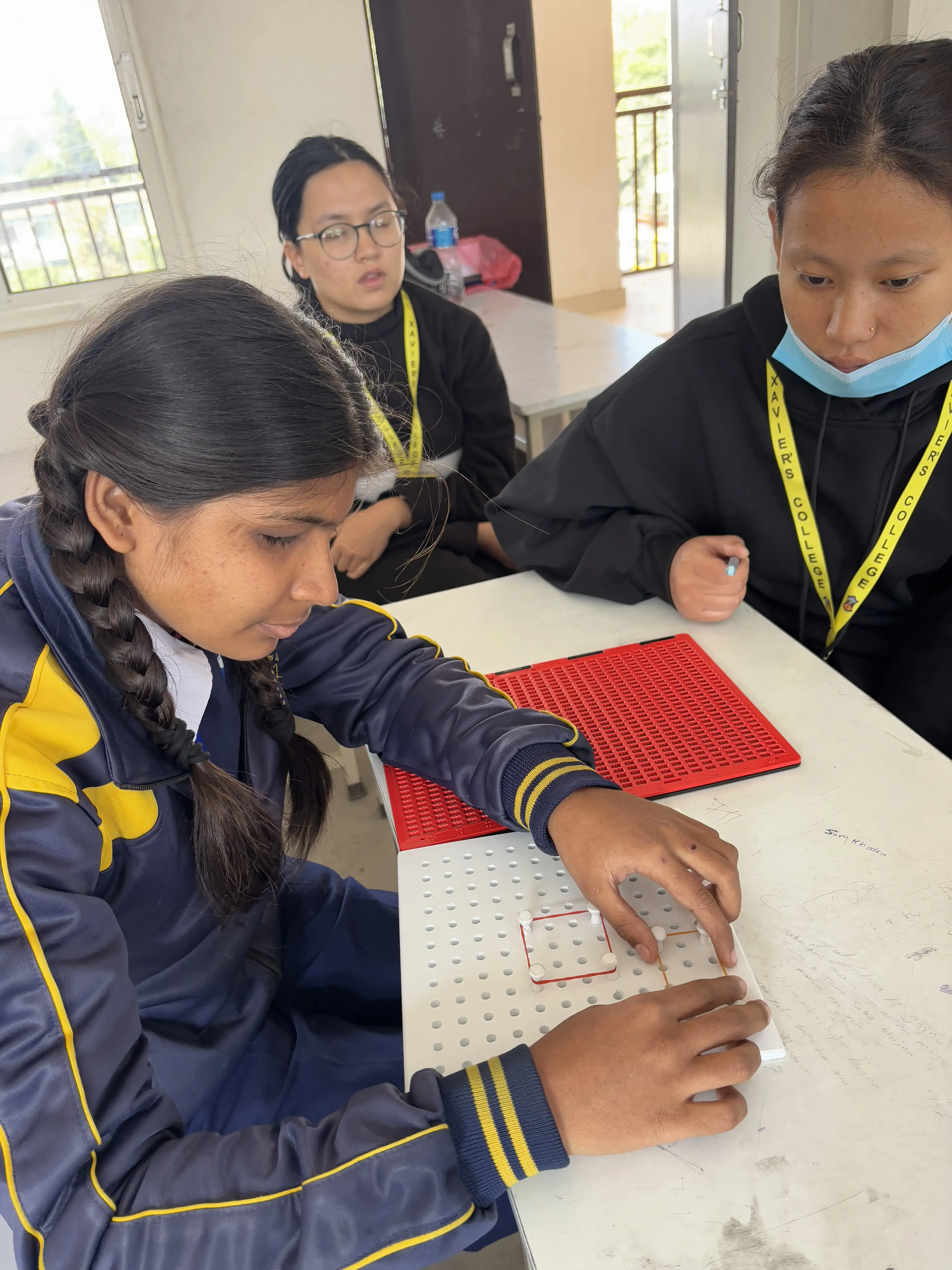 A picture where a female student with visual disability is interacting with 3D geometrical board and project member, Pushpa Rai is supporting her. In the background, Sanskriti Ghimire is also observed.