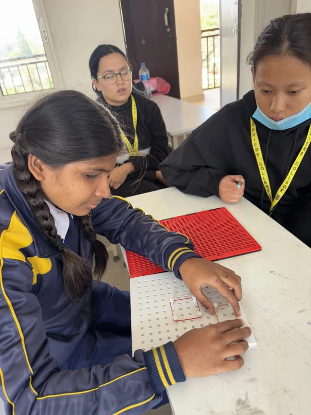 A picture where a female student with visual disability is interacting with 3D geometrical board and project member, Pushpa Rai is supporting her. In the background, Sanskriti Ghimire is also observed.