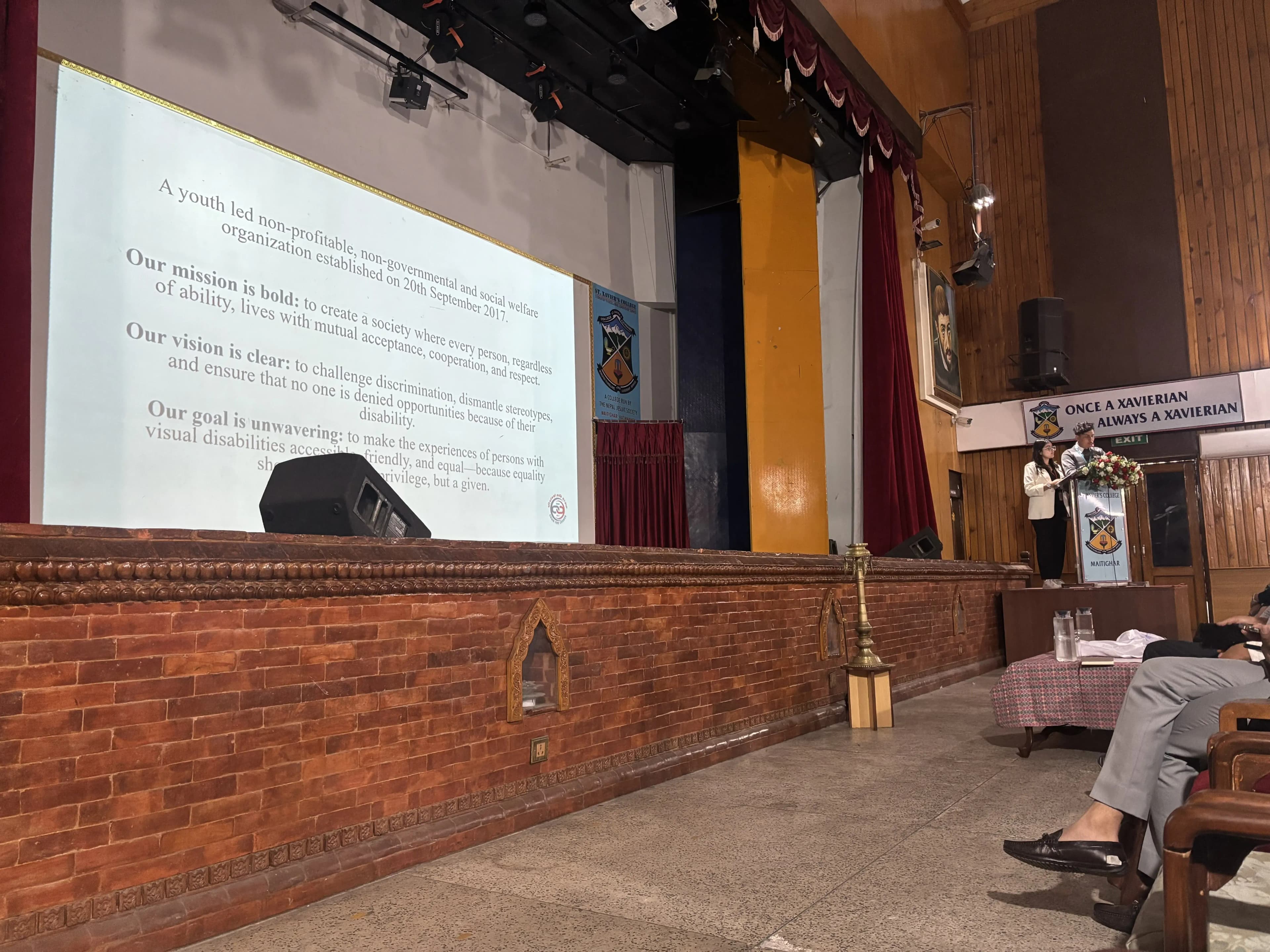 Our Public Relation & Philanthropy Officer, Mr. Sangam Khadka and Design Support Officer, Ms. Khushi Subedi, presenting about Project Wings to Dreams from the podium. In the background, a slide is being projected.
