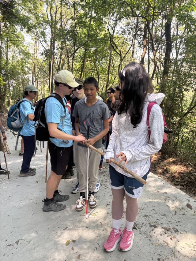 Two of the pair guides are holding the long stick in either sides and a participant with visual disability is holding it in between. One of the Spanish volunteers is explaining the participant with visual disability from side. In the background, other fellow hikers can be observed.