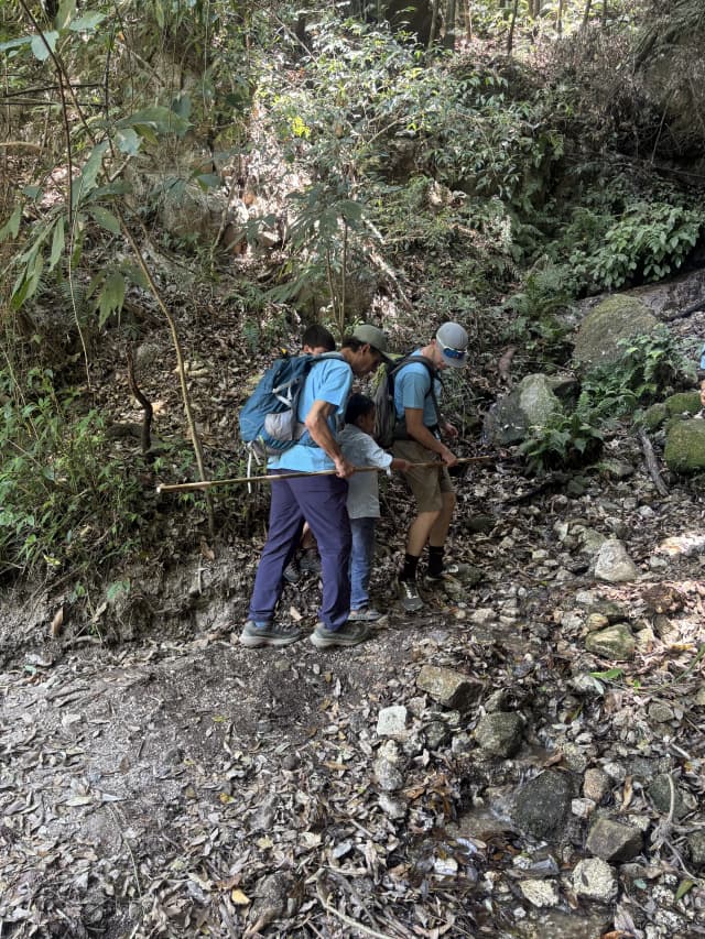 Two of the pair guides are holding the long stick in either sides and a participant with visual disability is holding it in between. In the background, green grasses and rocks can be observed.