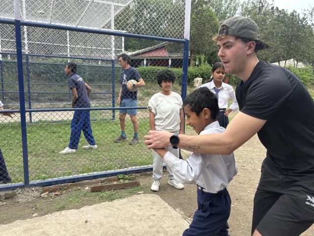 A Spanish intern is teaching one of the students with visual disabilities to throw the ball. In the background some of them are observing them and other playing stations are also observed.