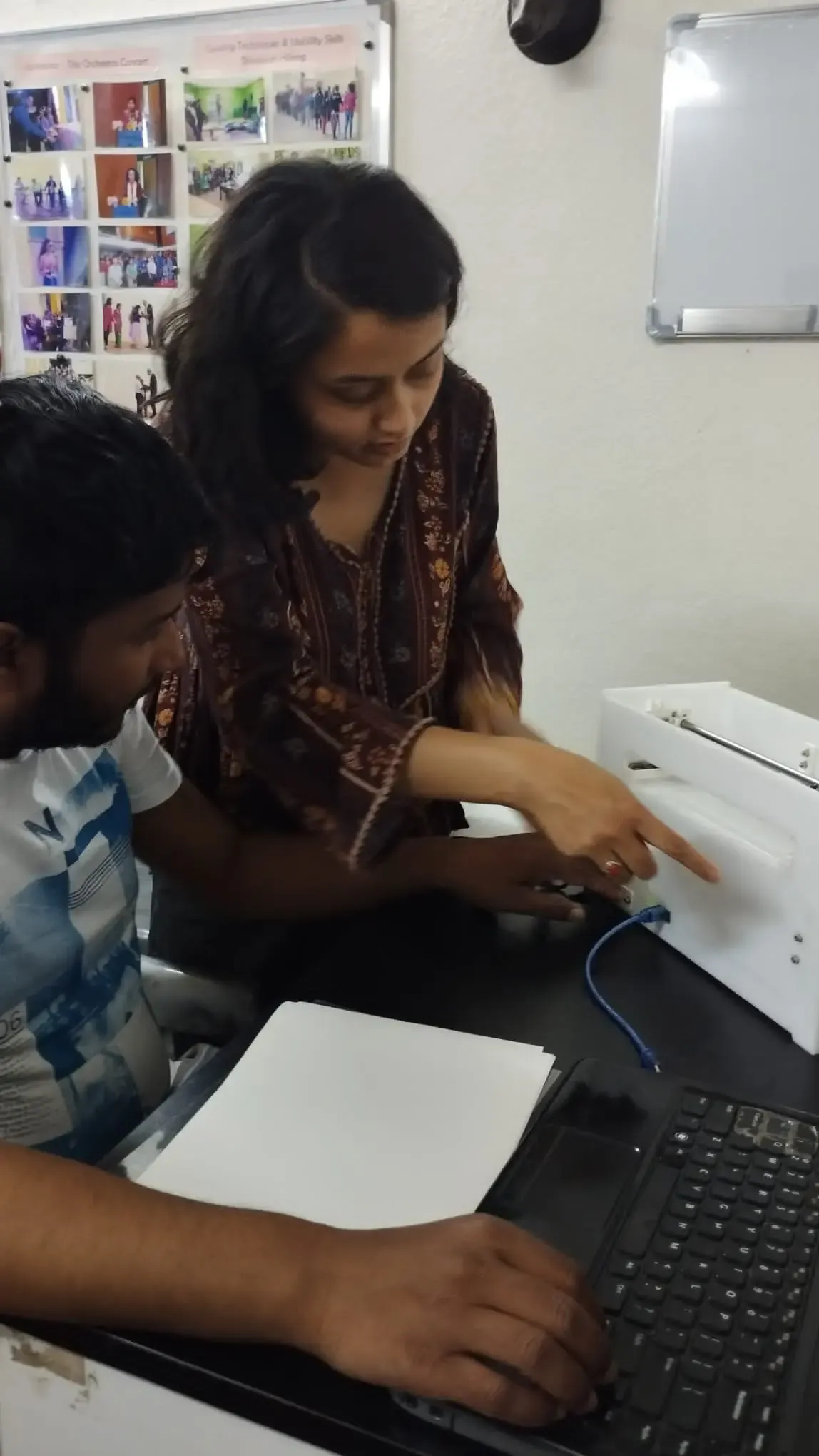 Secretary of CFC, Ms. Deepika Shahi is explaining about the Braille printer to the Founder of CFC, Mr. Abhishek Shahi. The Founder is touching the braille printer by one hand and with another hand he is using a laptop.