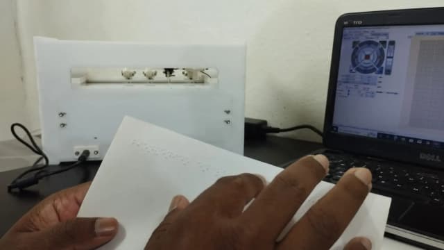 Our Founder, Mr. Abhishek Shahi is touching the printed braille paper. In the background, a braille printer and a laptop can be observed.