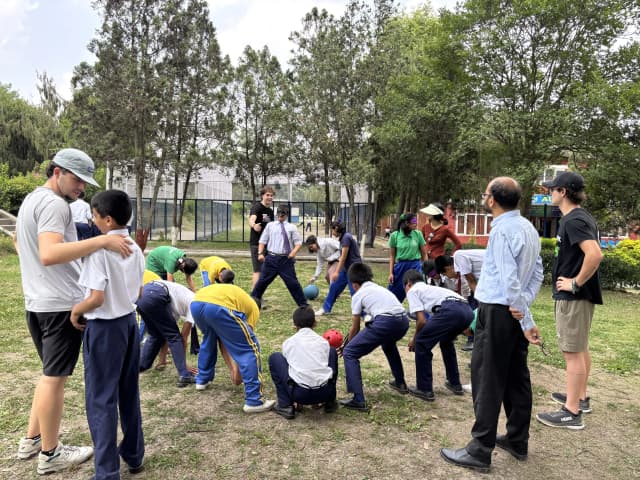 An image where children with visual disabilities are standing making a circle, playing a sound ball. Even Spanish interns are observed in the picture.