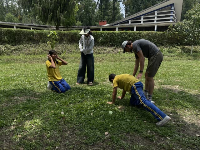 A male participant with visual disability is learning the defense position to block a ball and one of the Spanish interns is guiding him. A girl participant with visual disability and a project member are observing.