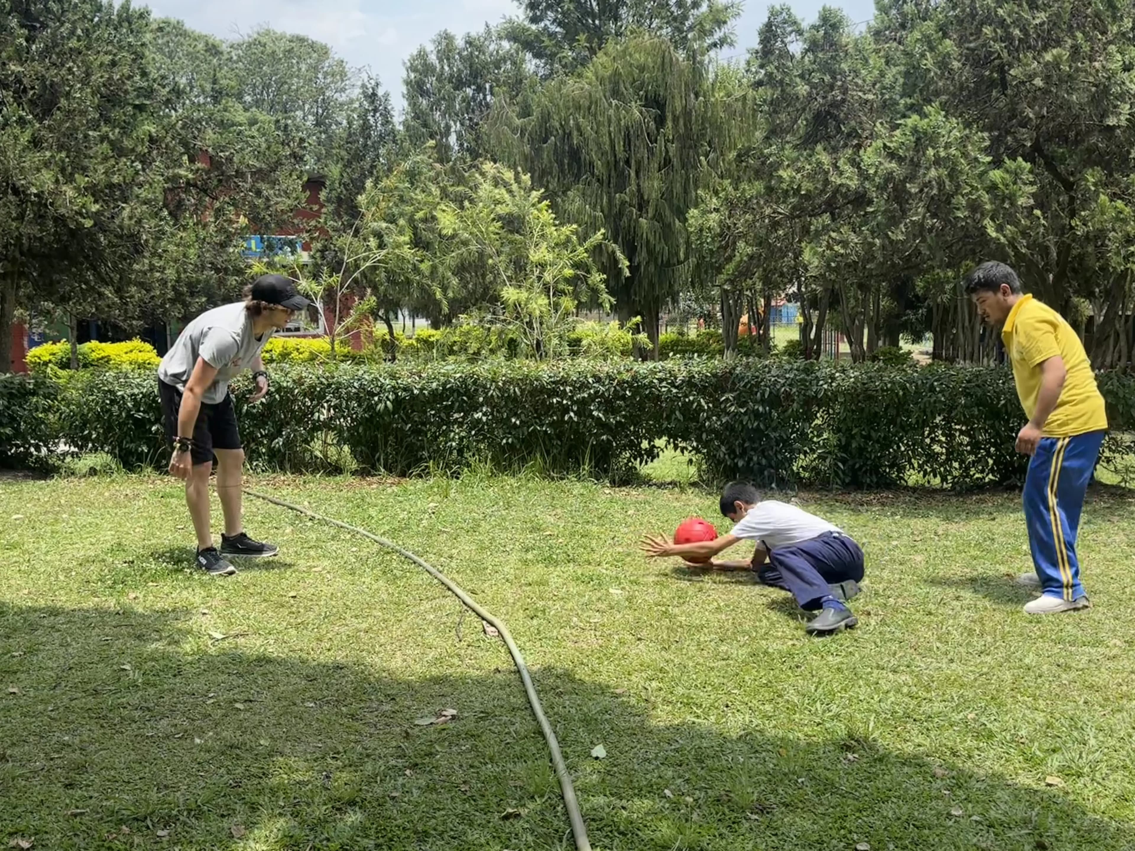 One of the Spanish interns is throwing a sound ball and a male participant with visual disability is catching it. Another male participant with visual disability is observe at the back.