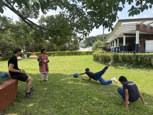 A male participant with visual disability is blocking a sound ball. His fellow partner is also observed. One of the Spanish interns and a female teacher is observing them.