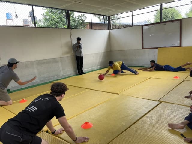 A team of two Spanish interns and two male participants with visual disabilities are playing goalball. One of the male participants with visual disability is trying to block the sound ball. In a background, one of the project member is also observed.