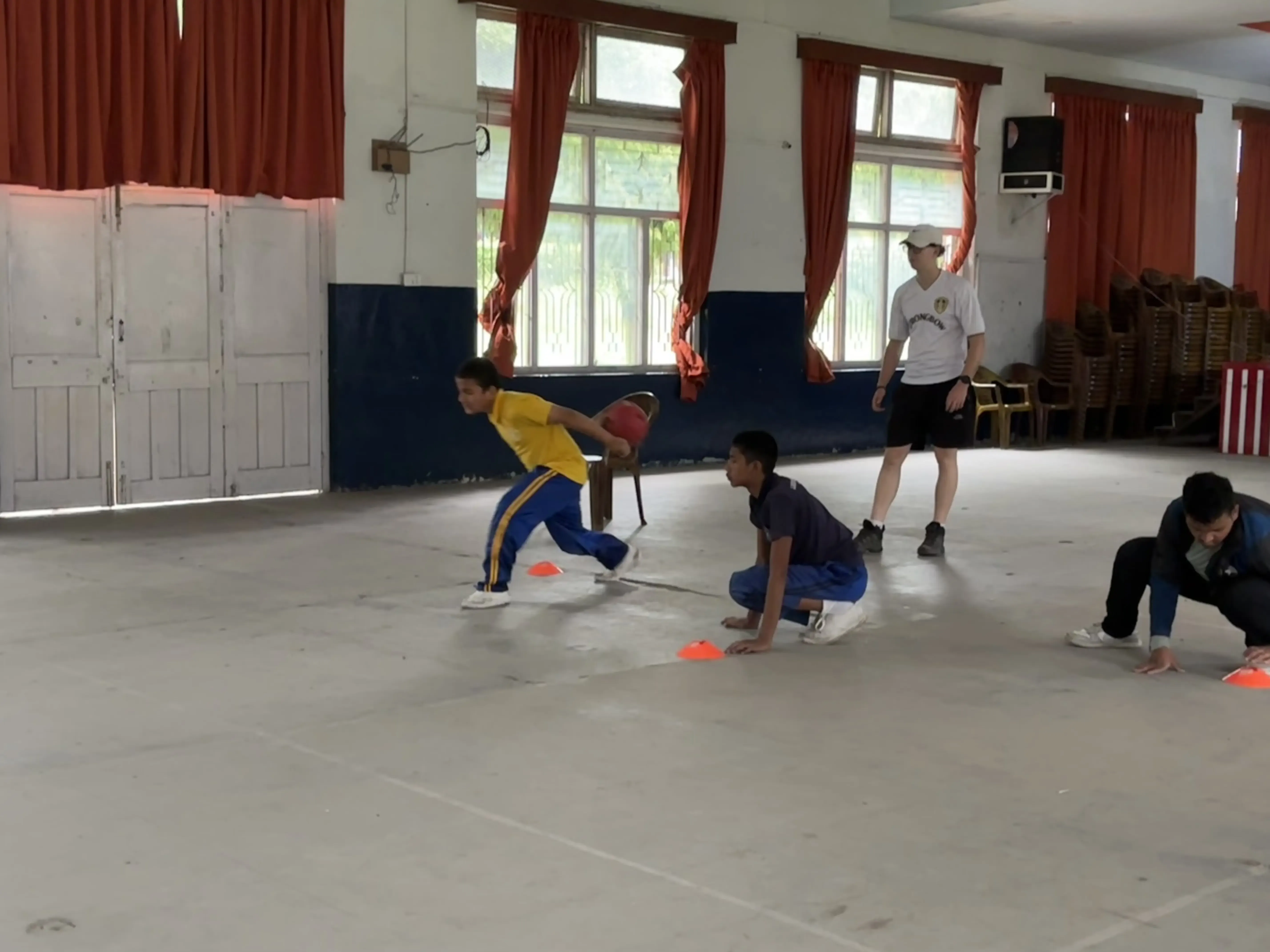 A team of 3 participants with visual disabilities are in their position playing goalball. One of them is throwing the sound ball. In the background, one of the Spanish interns is observed.