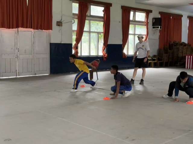 A team of 3 participants with visual disabilities are in their position playing goalball. One of them is throwing the sound ball. In the background, one of the Spanish interns is observed.