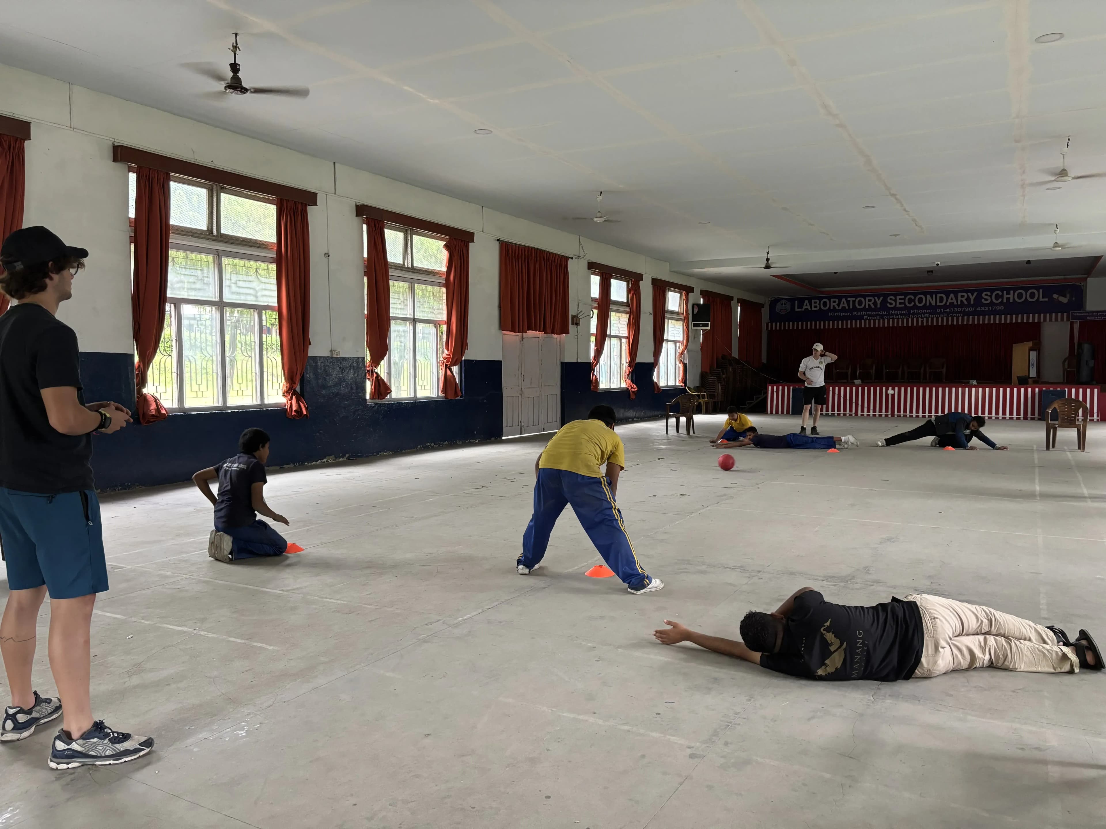 A full view of goalball court where 3 players with visual disabilities are defending their goalpost at either of the sides. The sound ball is rolling in between. On both the side, Spanish interns are standing and observing.