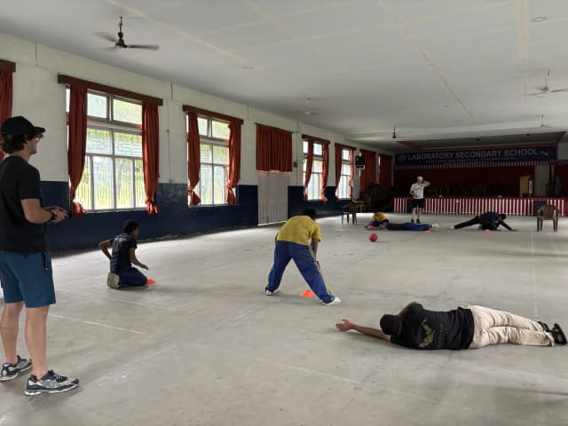 A full view of goalball court where 3 players with visual disabilities are defending their goalpost at either of the sides. The sound ball is rolling in between. On both the side, Spanish interns are standing and observing.