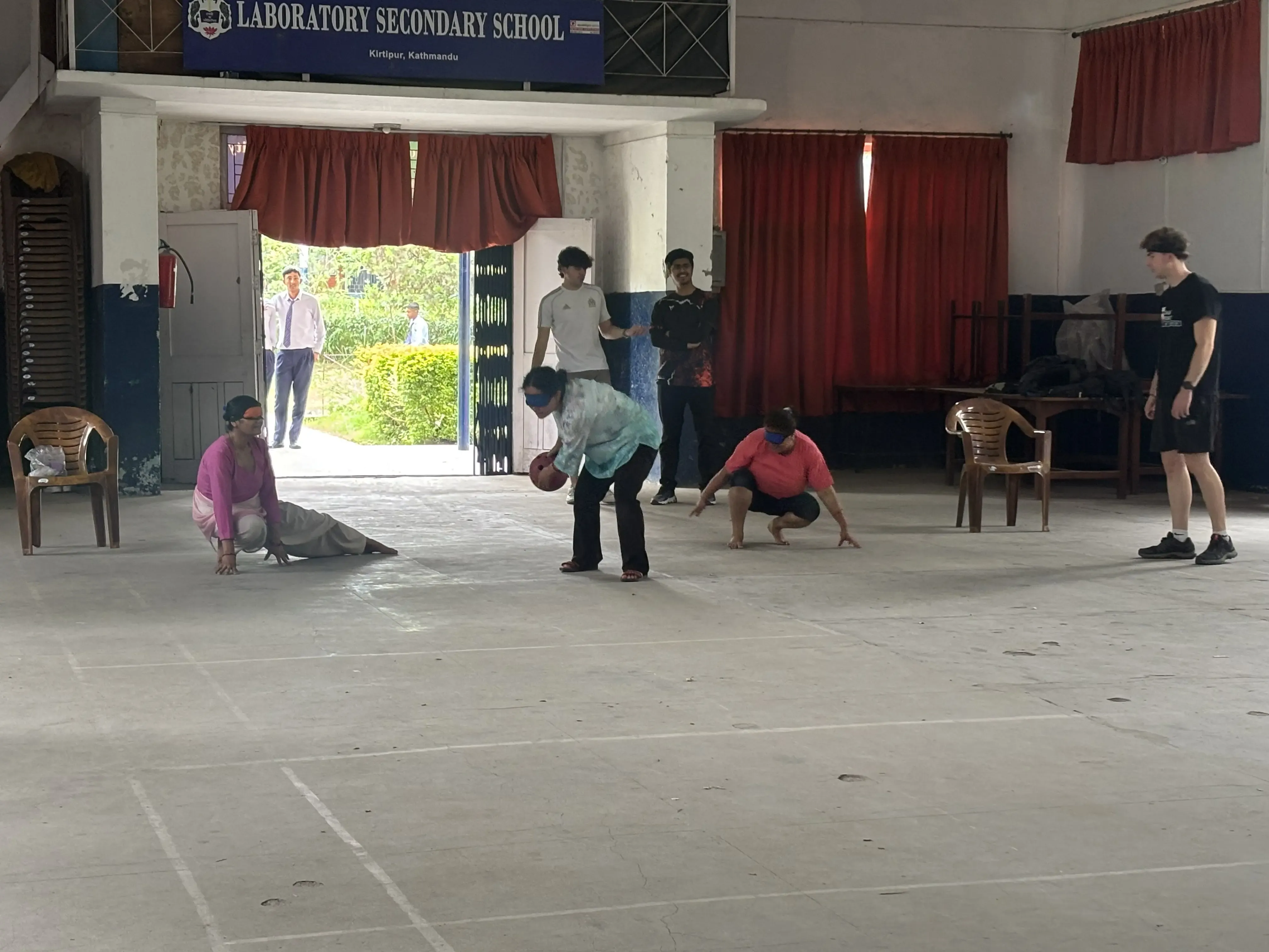 One of the members from teacher’s team is throwing a sound ball. All the 3 teachers are blindfolded. In the background, 2 of the Spanish interns and a project member is observed.