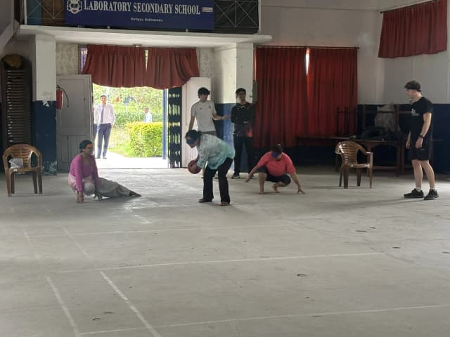 One of the members from teacher’s team is throwing a sound ball. All the 3 teachers are blindfolded. In the background, 2 of the Spanish interns and a project member is observed.