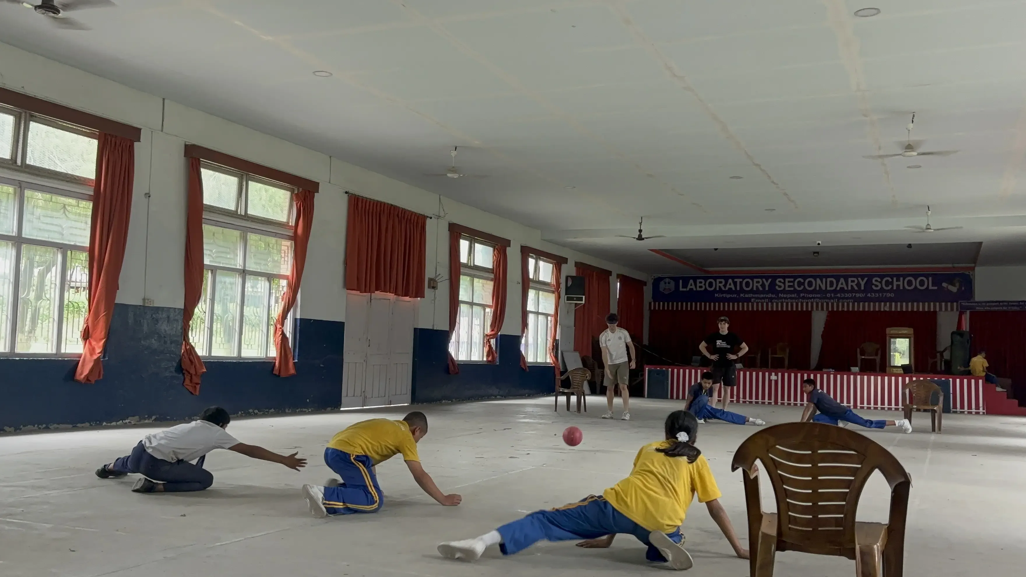 A full view of goalball court where 3 players with visual disabilities are defending their goalpost at one of the side. In opposing side, a Spanish intern who is a part of the team is throwing the sound ball and his fellow team members are taking their position. One of the Spanish interns is standing and observing.
