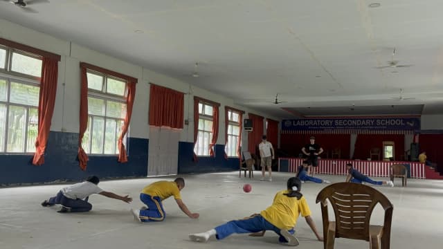A full view of goalball court where 3 players with visual disabilities are defending their goalpost at one of the side. In opposing side, a Spanish intern who is a part of the team is throwing the sound ball and his fellow team members are taking their position. One of the Spanish interns is standing and observing.