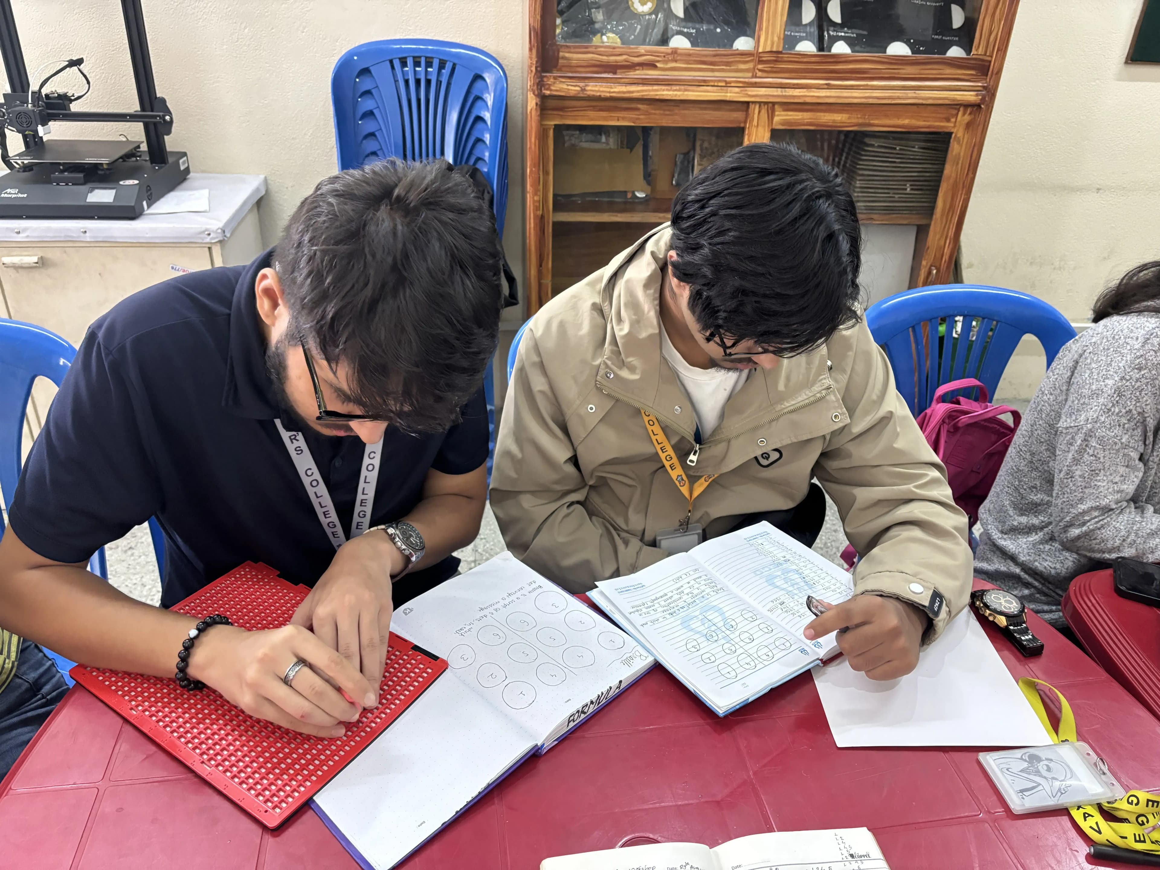 One of the male participants is looking at his notebook and writing the braille. A fellow participant is looking at his note.
