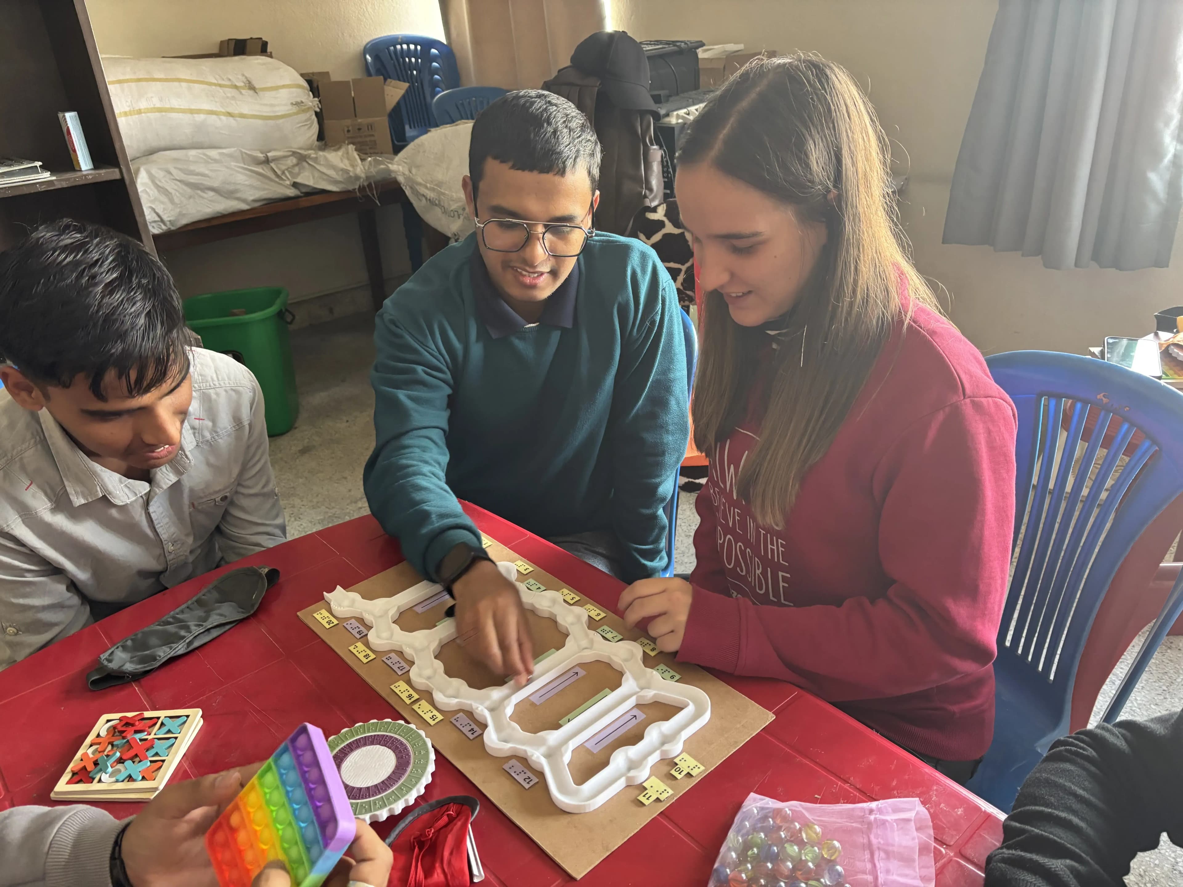 Three people, two men and a woman, are seated around a red table engaged in an activity involving tactile educational materials. The woman is wearing a red sweatshirt, and the man in the middle has glasses and a green sweater. They are interacting with a board that appears to have raised or 3D paths, possibly for a learning or sensory game, and labeled sections with Braille and printed text. On the table are other tactile or sensory items, such as a rainbow-colored pop-it toy, a tic-tac-toe puzzle, and a bag of marbles.