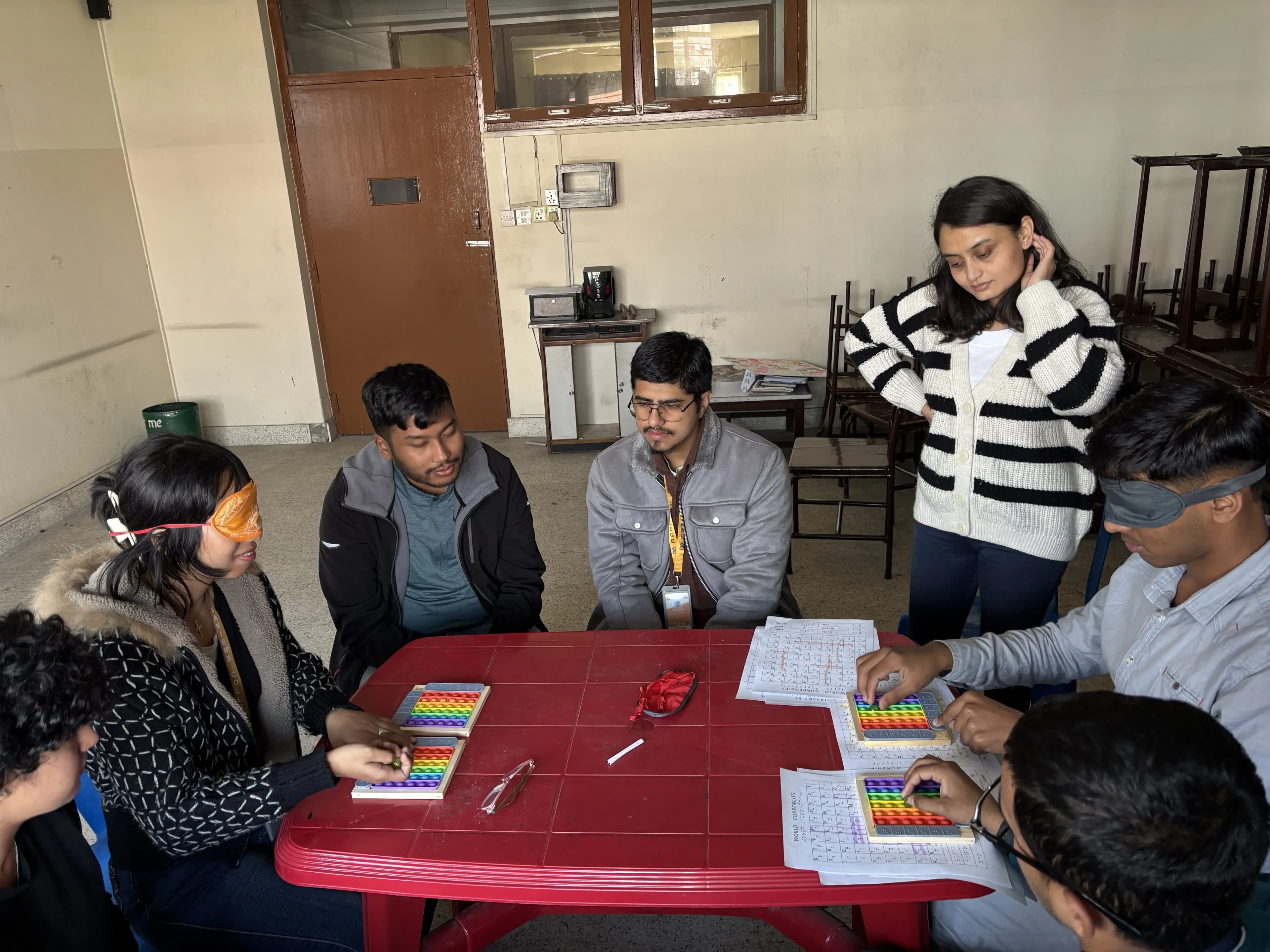 Group of six people sitting around a rectangular red table in a room. Several of them are using colorful rainbow pop-it fidget toys and are also holding or working on sheets of paper with printed or possibly Braille text. Some of the participants are blindfolded, suggesting an activity focused on sensory or tactile experience, possibly to simulate visual impairment. The mood is collaborative and focused. The background includes stacked chairs, a bed, a brown door, and some visible electrical fixtures.
