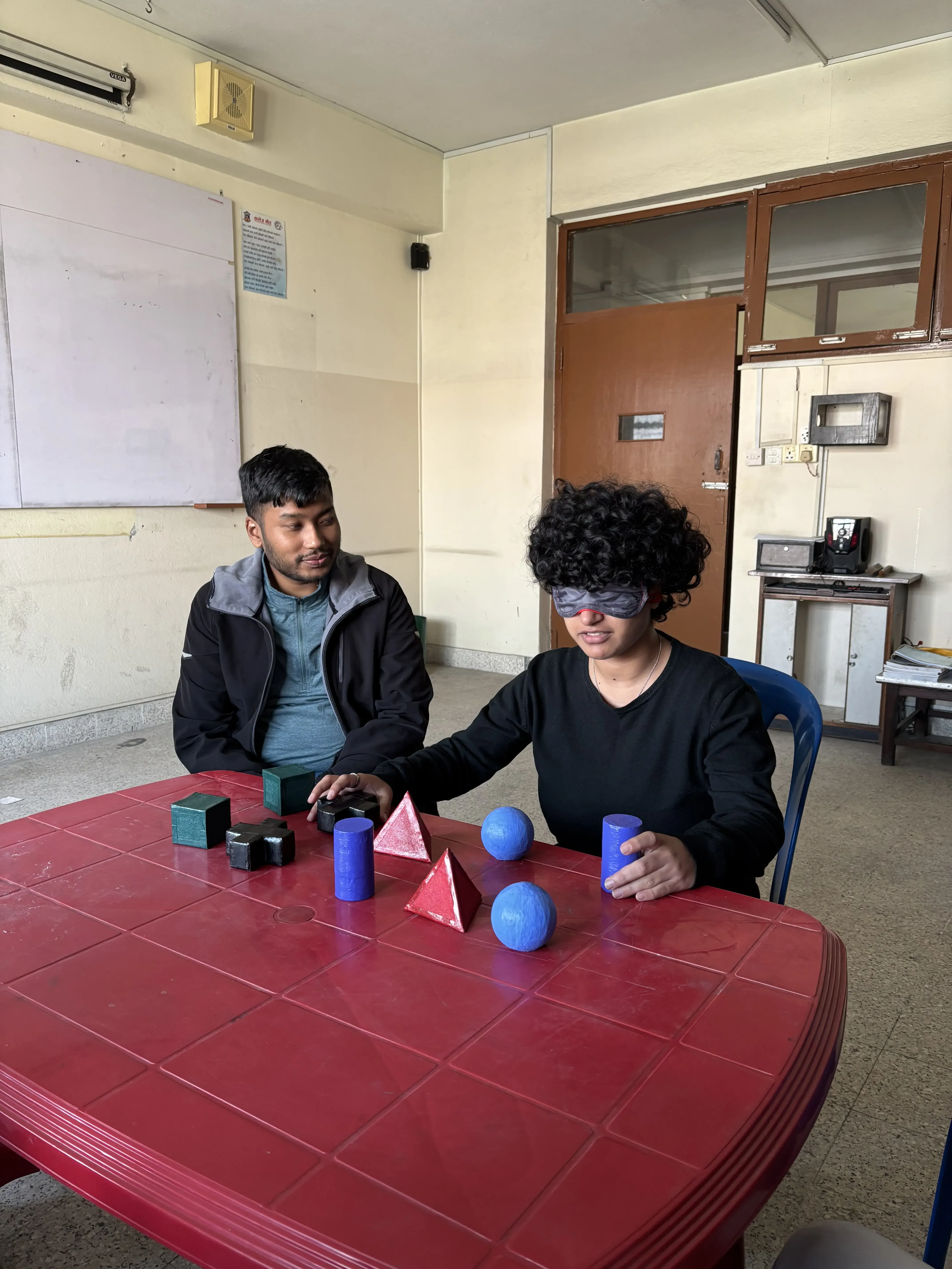 Two men sitting at a red table in a room. One man has curly hair and is wearing a blindfold while interacting with various colorful 3D geometric shapes on the table, such as cones, spheres, cubes, cylinders, and pyramids in blue, red, green, and black. The other man is sitting next to him, watching the activity and smiling. The background shows a brown door, a glass window, a whiteboard on the wall, and some classroom or office items.