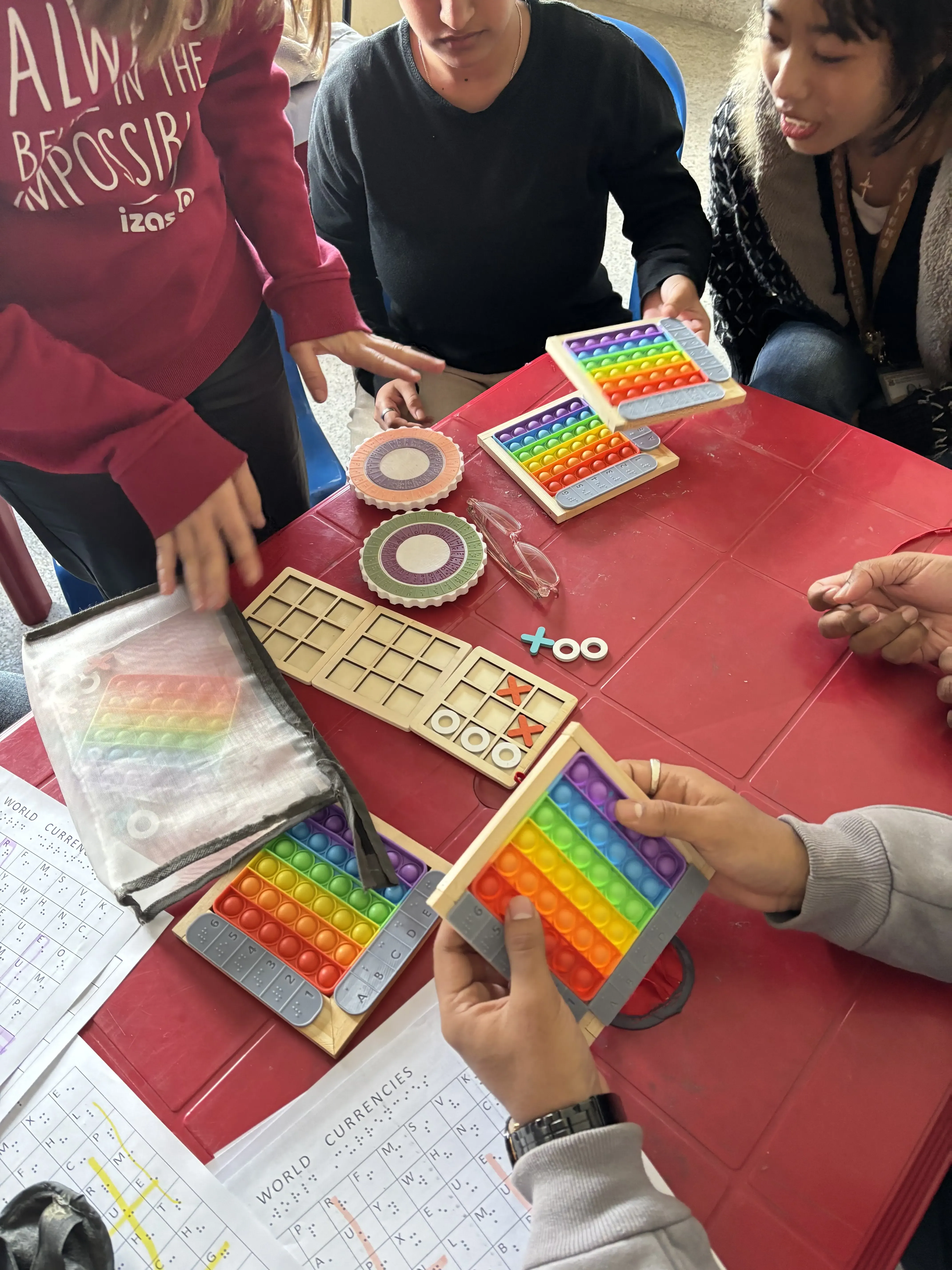 Red table covered with colorful sensory and educational materials. Several people are seated or standing around it, with their hands visible engaging with the items. On the table are multiple rainbow-colored pop-it fidget toys, two tactile tic-tac-toe boards with X and O pieces, two round tactile discs, a mesh bag containing another pop-it toy, and several sheets of paper with word searches or puzzles titled WORLD CURRENCIES.
