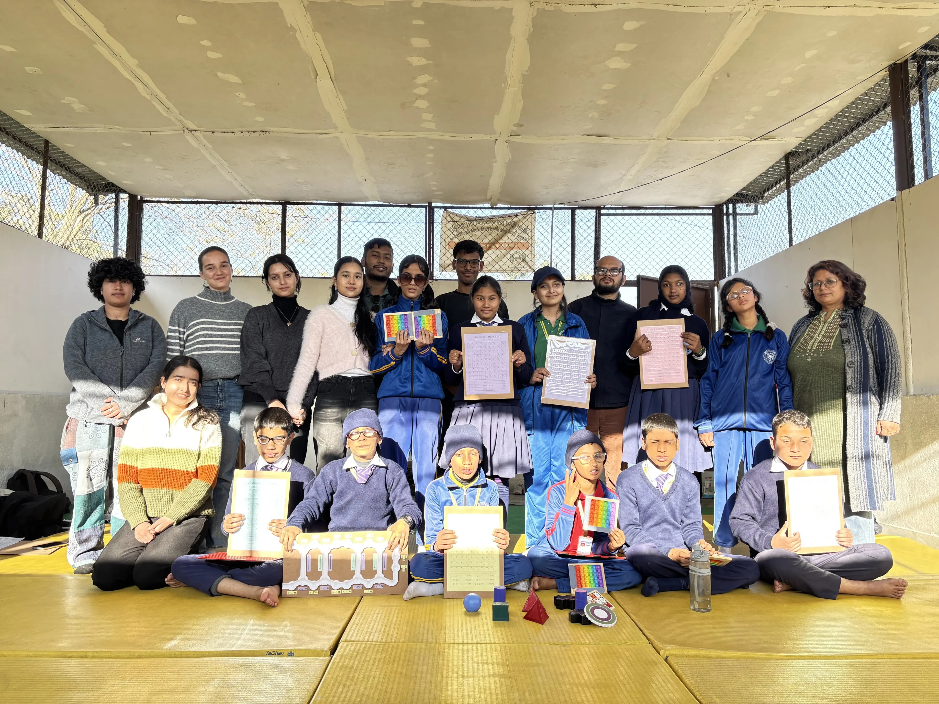 The picture shows a large group of people, including adults and children wearing school uniforms, posing together in what appears to be an open indoor space with yellow flooring and walls with fencing. Several individuals are holding colorful tactile learning materials such as pop-it toys, geometric shapes, and word puzzles.