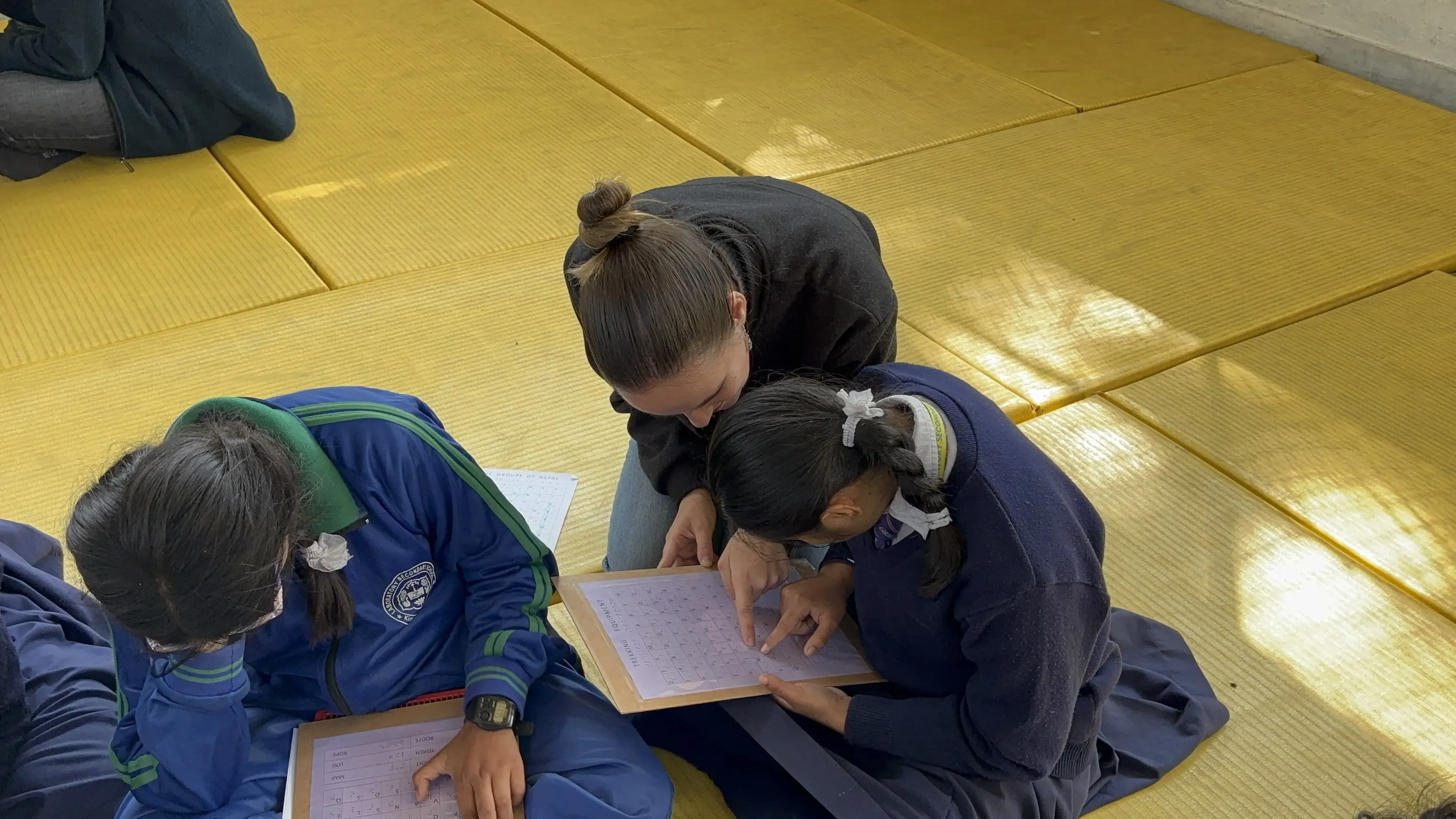 Two schoolgirls and an adult are seated on a yellow padded mat, focused on a tactile worksheet. The adult, who has hair tied in a bun and is wearing a dark top, is leaning in to assist one of the girls, who is using her finger to trace or read the worksheet—most likely a word search similar to those seen earlier. The girls are dressed in school uniforms: one in a blue tracksuit with green trim and the school crest, the other in a blue sweater and skirt with a white shirt and tie.