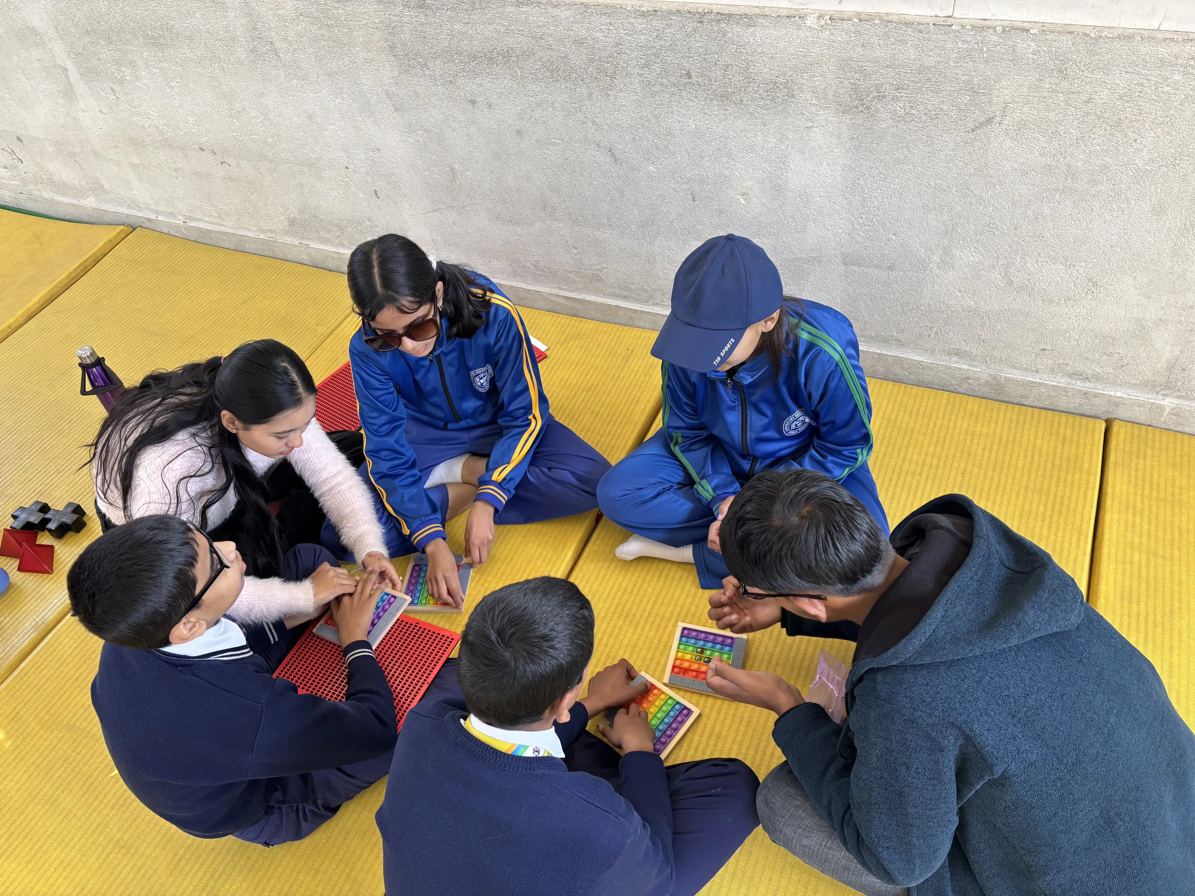 A group of seven students is seated in a circle on yellow padded mats in front of a light-colored wall. They are engaged with tactile learning materials, including colorful pop-it fidget toys and other tactile items. Most of the students are wearing school uniforms, with some in blue tracksuits with green accents, and a couple of students in dark sweaters.