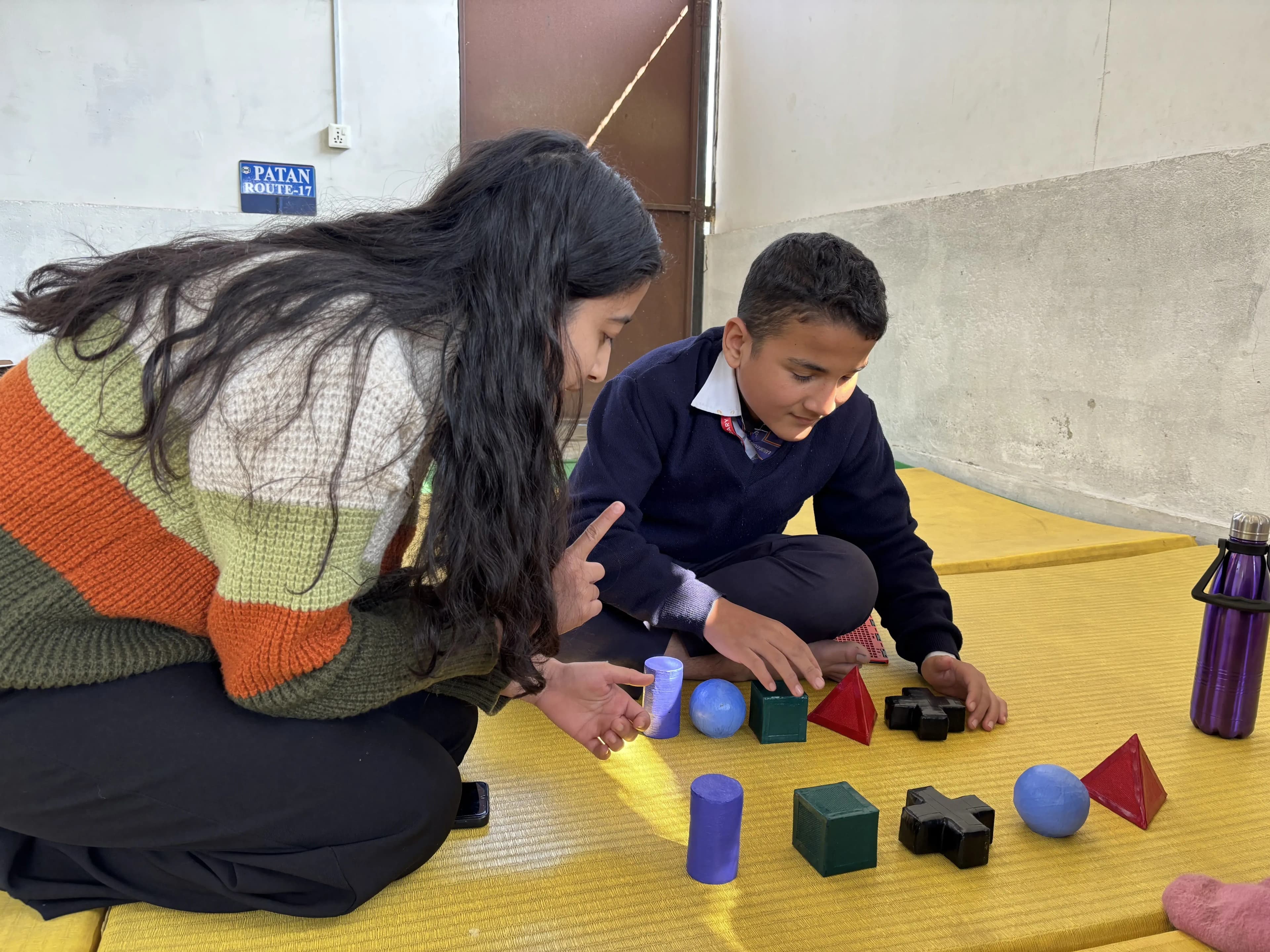 A woman and a young boy are sitting on yellow padded mats in front of a light-colored wall, engaging with a variety of colorful 3D geometric shapes spread out in front of them. The woman is wearing a green, orange, and cream striped sweater and is interacting attentively with the boy, who is dressed in a navy school uniform with a white shirt. They both appear focused on the tactile objects, suggesting a hands-on learning or teaching moment.