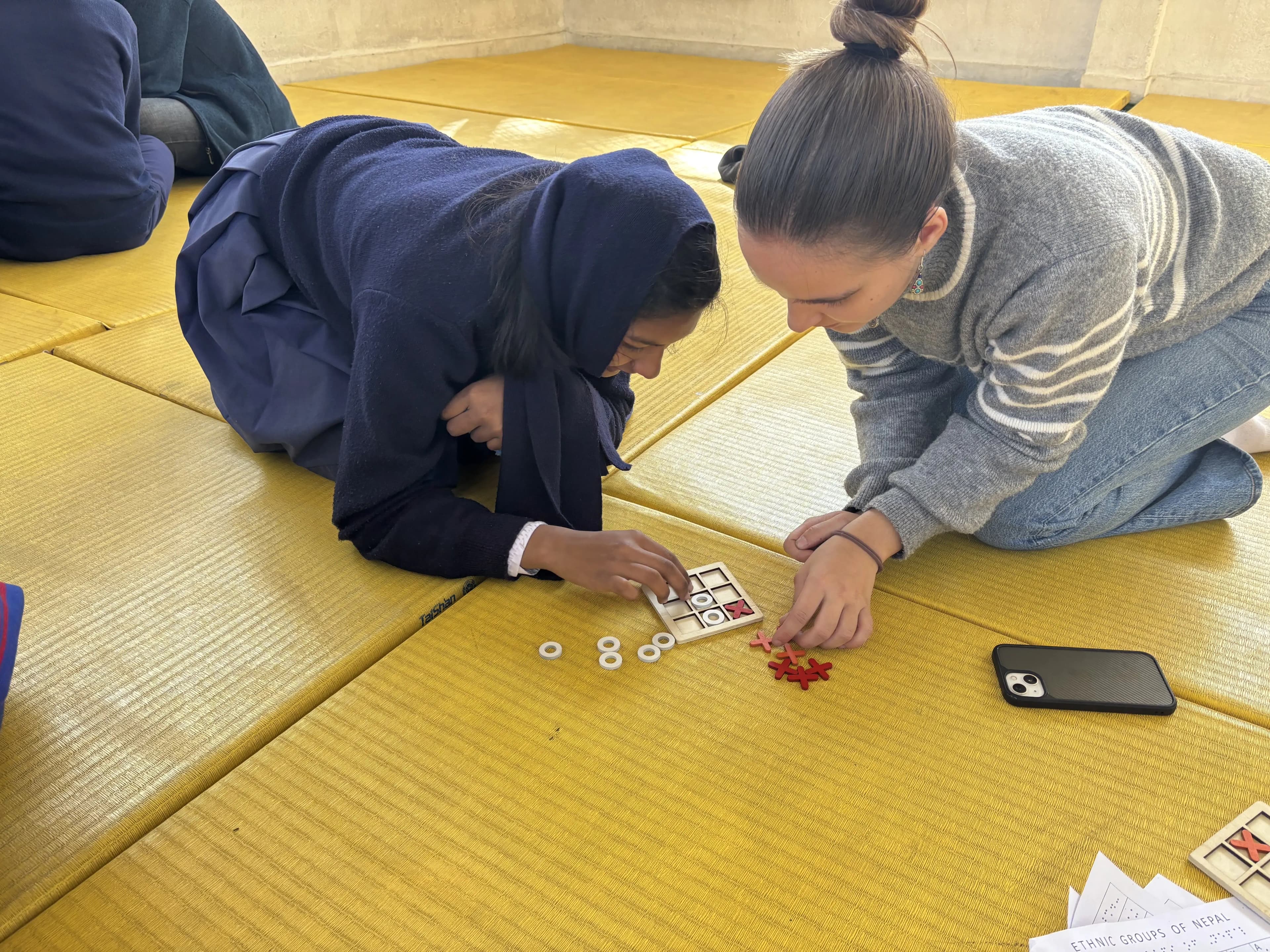 A girl in a dark blue school uniform and a woman in a light gray sweater with her hair in a bun are sitting on yellow padded mats. They are both focused on a tactile activity on the floor, which appears to be a tic-tac-toe game using a small wooden board and plastic X and O pieces. The woman is helping arrange the pieces while the girl is actively engaged in the game. A smartphone and some papers are placed nearby on the mat.