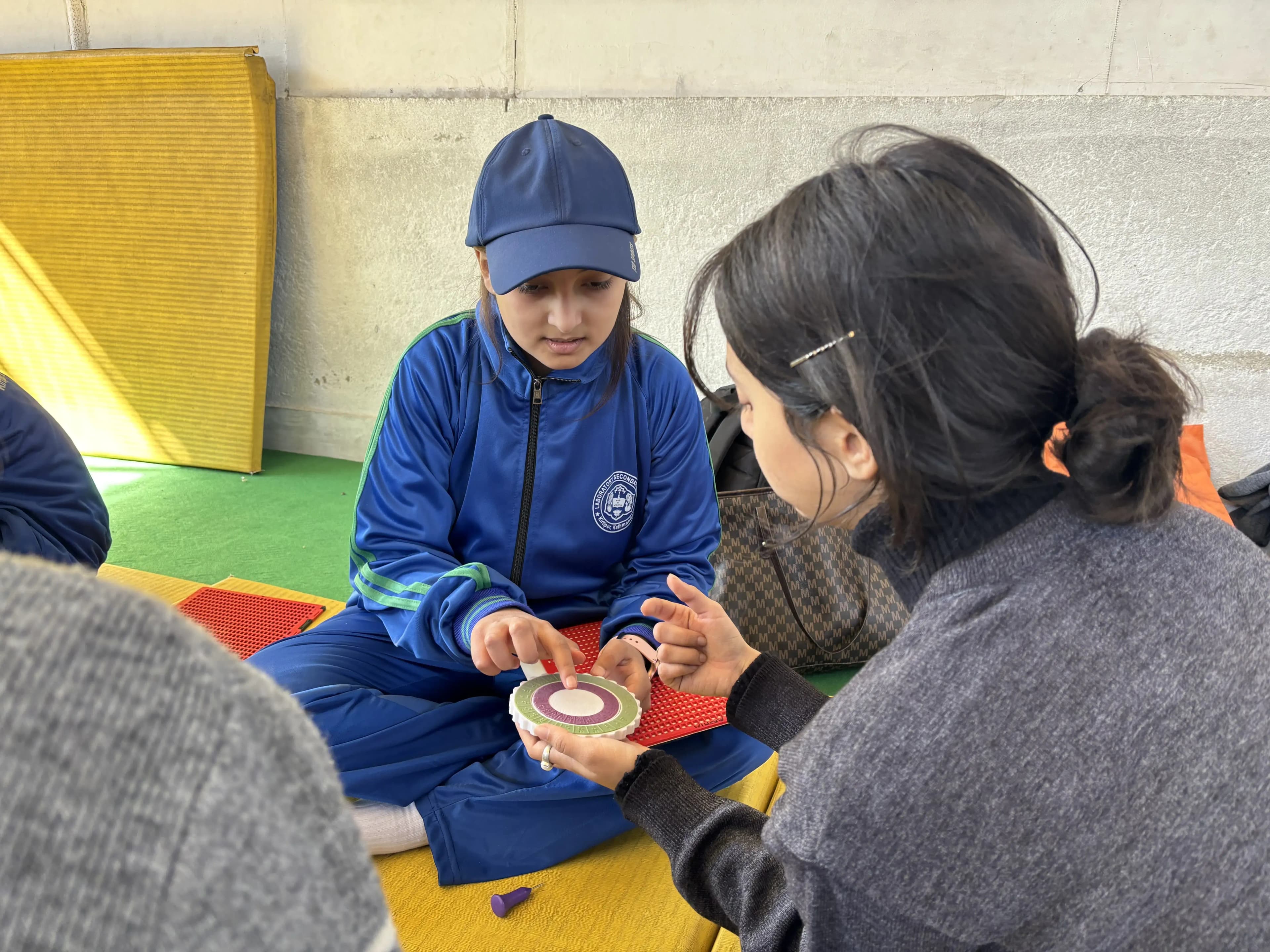 A group of people sitting on colorful foam mats against a light-colored wall. In the center, a child in a blue tracksuit and blue cap (likely a school uniform) is attentively touching a round tactile object, which is being held by an adult woman with dark hair tied back. The adult is guiding the child's hands as they explore the textured surface of the object, which looks like an adaptive learning or sensory tool. There are other tactile objects scattered around, and another child and adult are partially visible, focused on the activity.