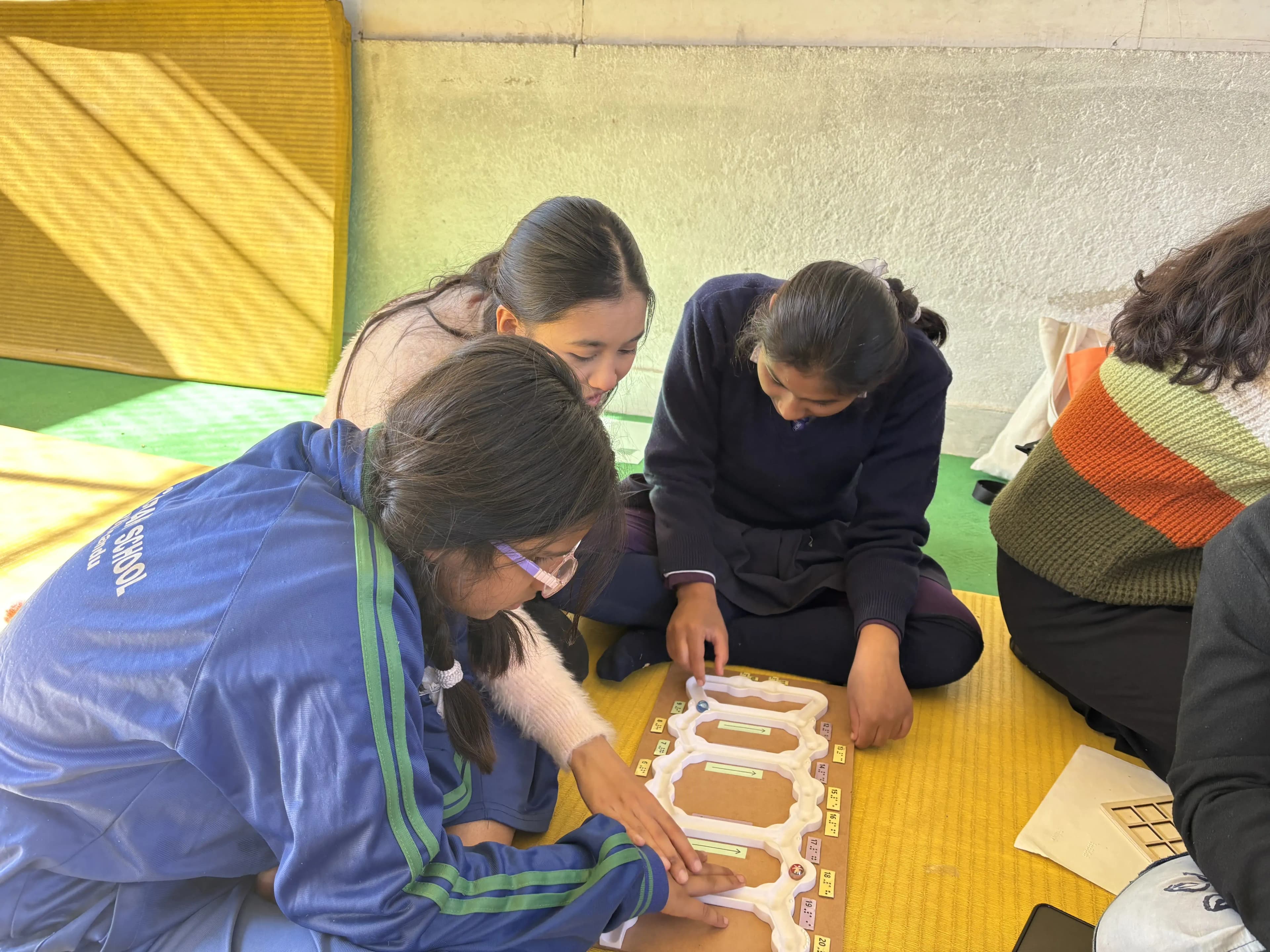 Four children and an adult are seated on colorful mats near a wall, focusing on a tactile educational board game with raised sections, possibly used for a sensory or adaptive learning activity. The children are in school uniforms, with one child wearing a blue tracksuit and others in navy sweaters. The adult, wearing a sweater with orange, green, and cream colors, supports and engages with the students as they interact with the tactile board.