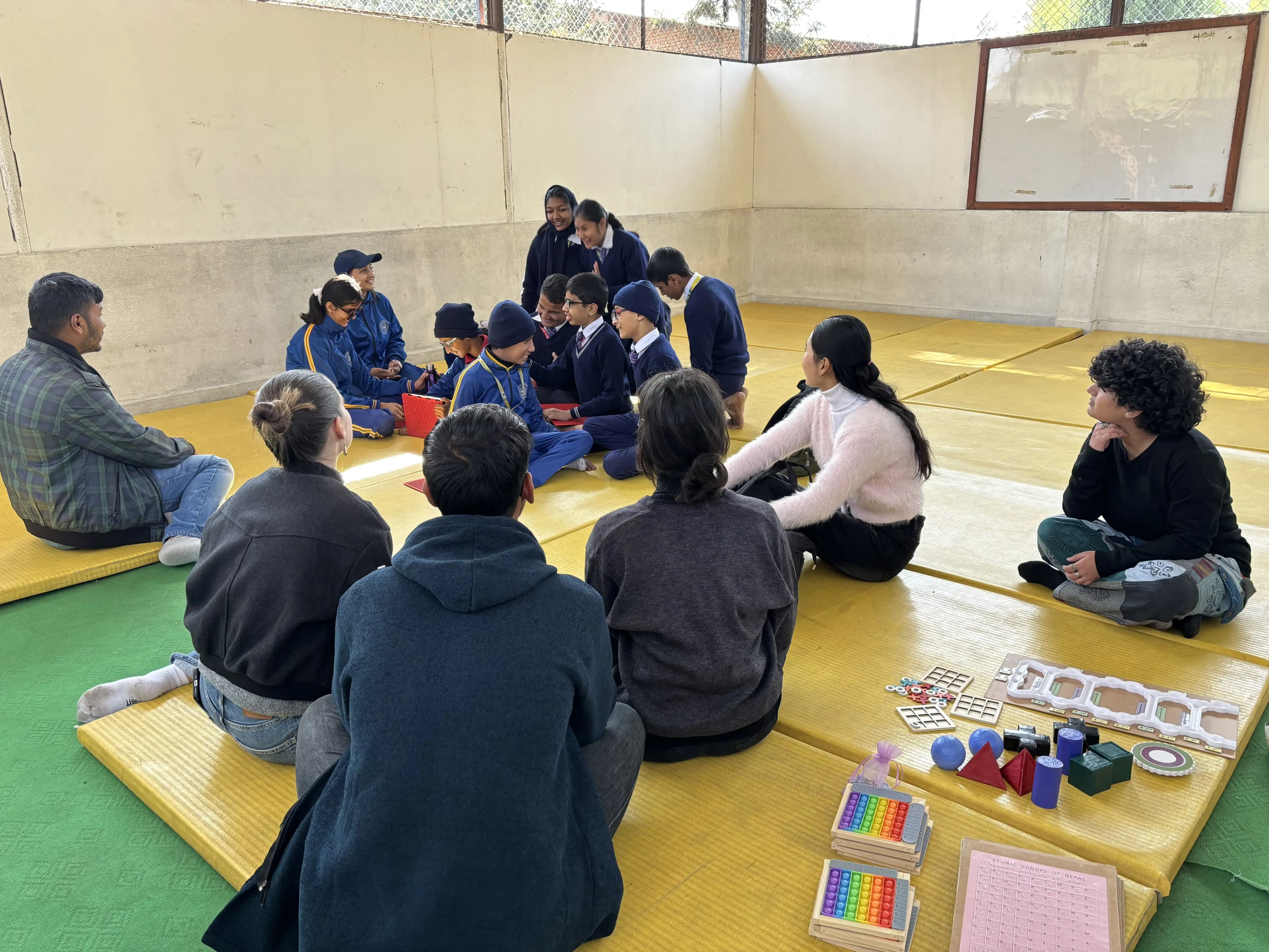 A group of students and adults are gathered in a spacious indoor area with yellow padded mats on the floor. Most participants are sitting in a large circle, and the focus appears to be on a central group of students who are engaged in an activity or discussion. The children, many of whom are wearing blue school uniforms or tracksuits, are interacting closely together, while the adults observe and facilitate from around the outside of the circle. On the mats near the camera, there are a variety of tactile and adaptive educational materials, such as rainbow-colored pop-it toys, geometric shapes, circular tactile boards, and word search sheets.