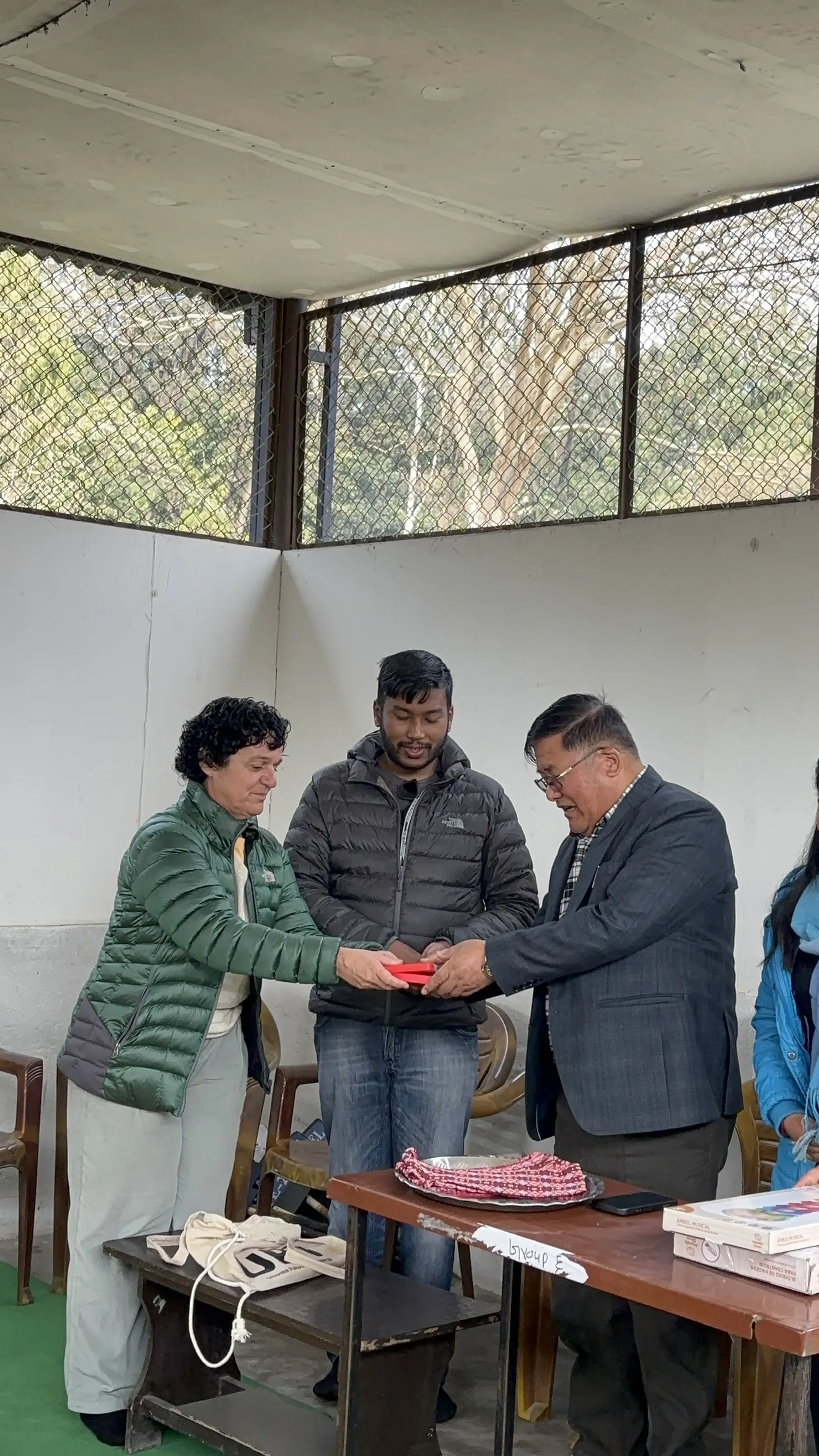 Three adults standing together indoors, with two of them (a man in a suit and a woman in a green jacket) holding a red object together. The man between them, in a grey jacket, is looking at the object and smiling. There are chairs, a small bench with drawstring bags, a table with boxes, and a tray with ropes or braided items nearby.