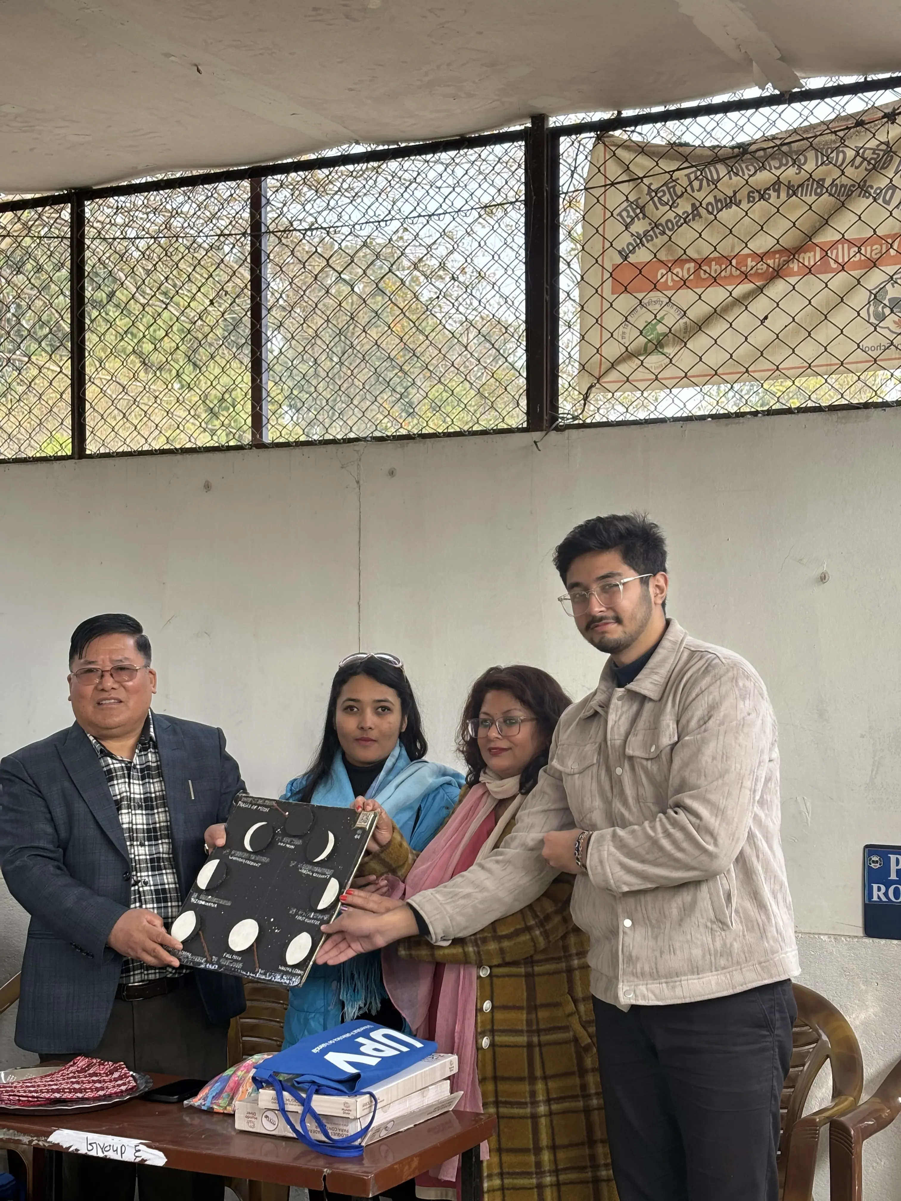 Four adults, one man and three women are indoors, standing next to a table and holding a black model that shows the phases of the Moon. They are taking part in a formal event to hand over the accessible 3D model to the representative of the school.