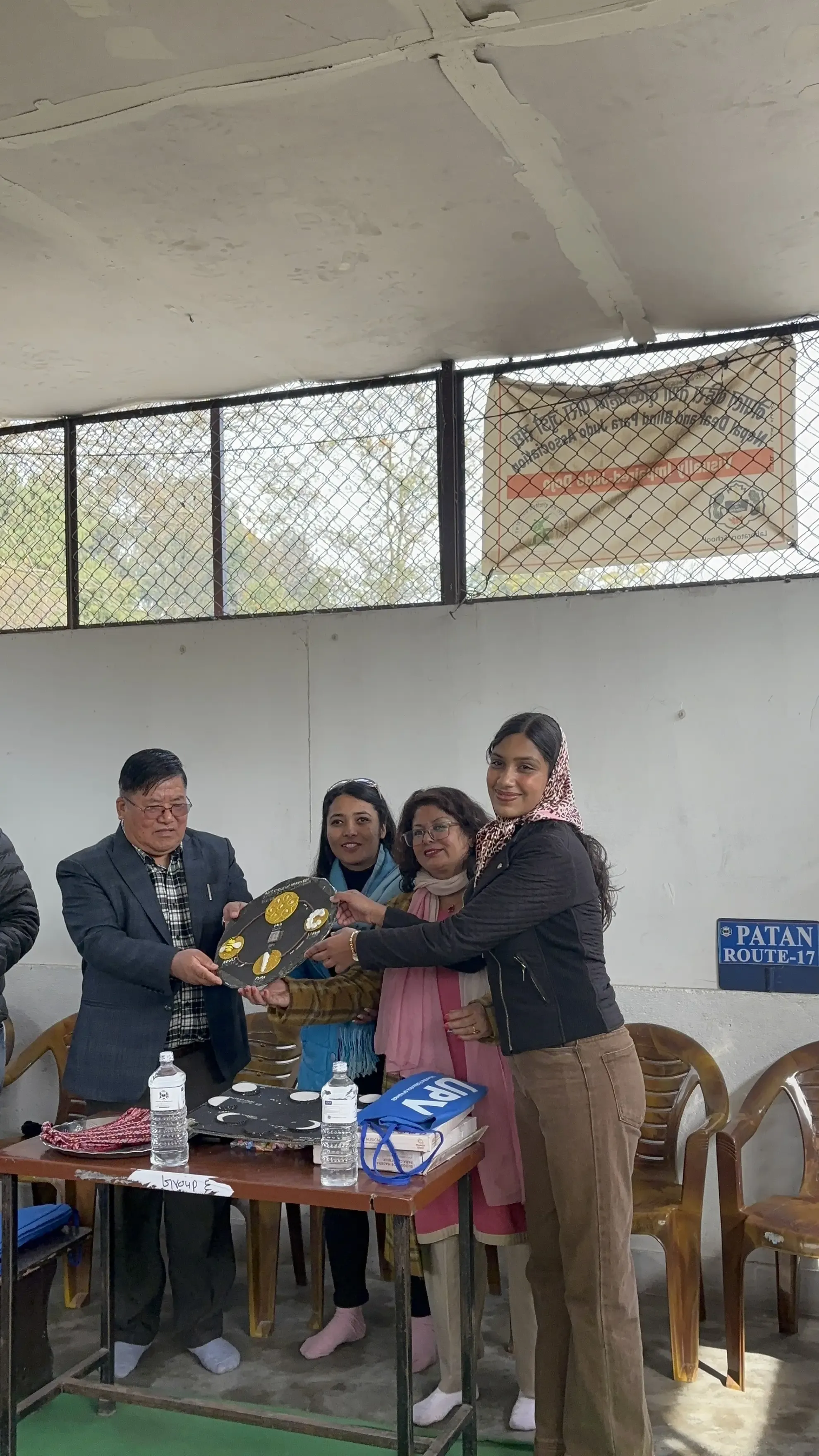 Four adults, one man and three women are standing indoors, gathered around a table. They are holding a black circular model that shows the lifecycle of a honeybee. The group is participating in a formal handover event of the accessible 3D model to the representative of the school.