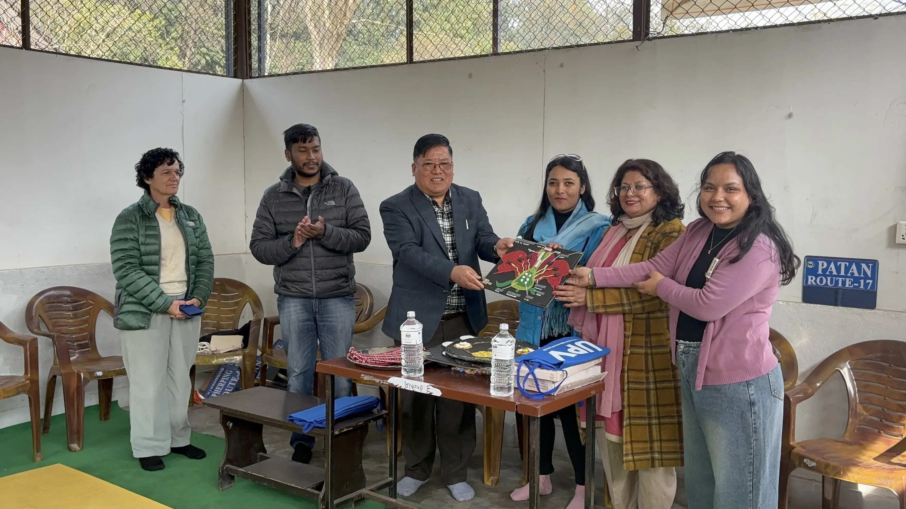 Six adults, including four women and two men, are standing together indoors near a table. The man in the suit and three women are holding a rectangular 3D educational model showing the parts of a flower. They are taking part in a formal event to hand over the accessible 3D model to the representative of the school. The atmosphere is positive, with some participants smiling and others applauding.