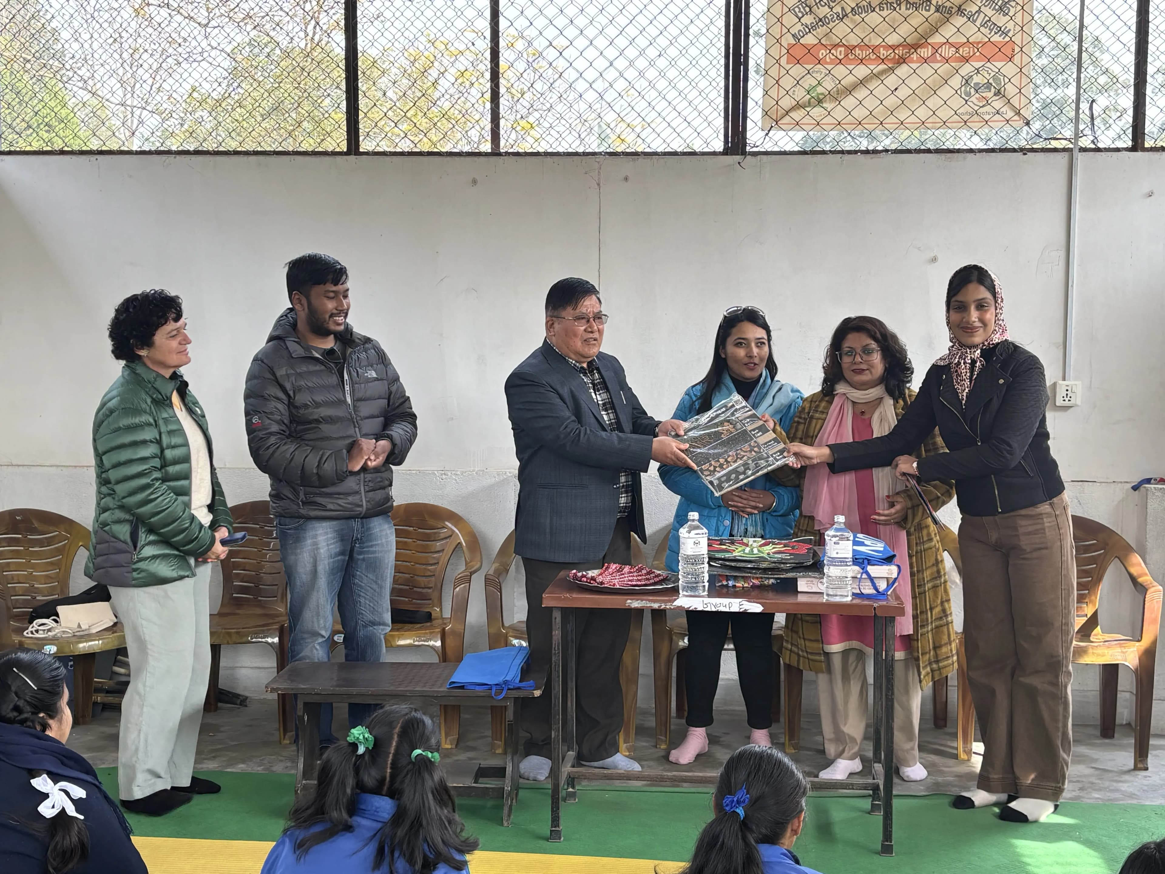 Six adults, including four women and two men, are standing together indoors near a table. The man in the suit and three women are holding a rectangular 3D educational model showing the layers of soil. They are taking part in a formal event to hand over the accessible 3D model to the representative of the school. The atmosphere is positive, with some participants smiling and others watching attentively.