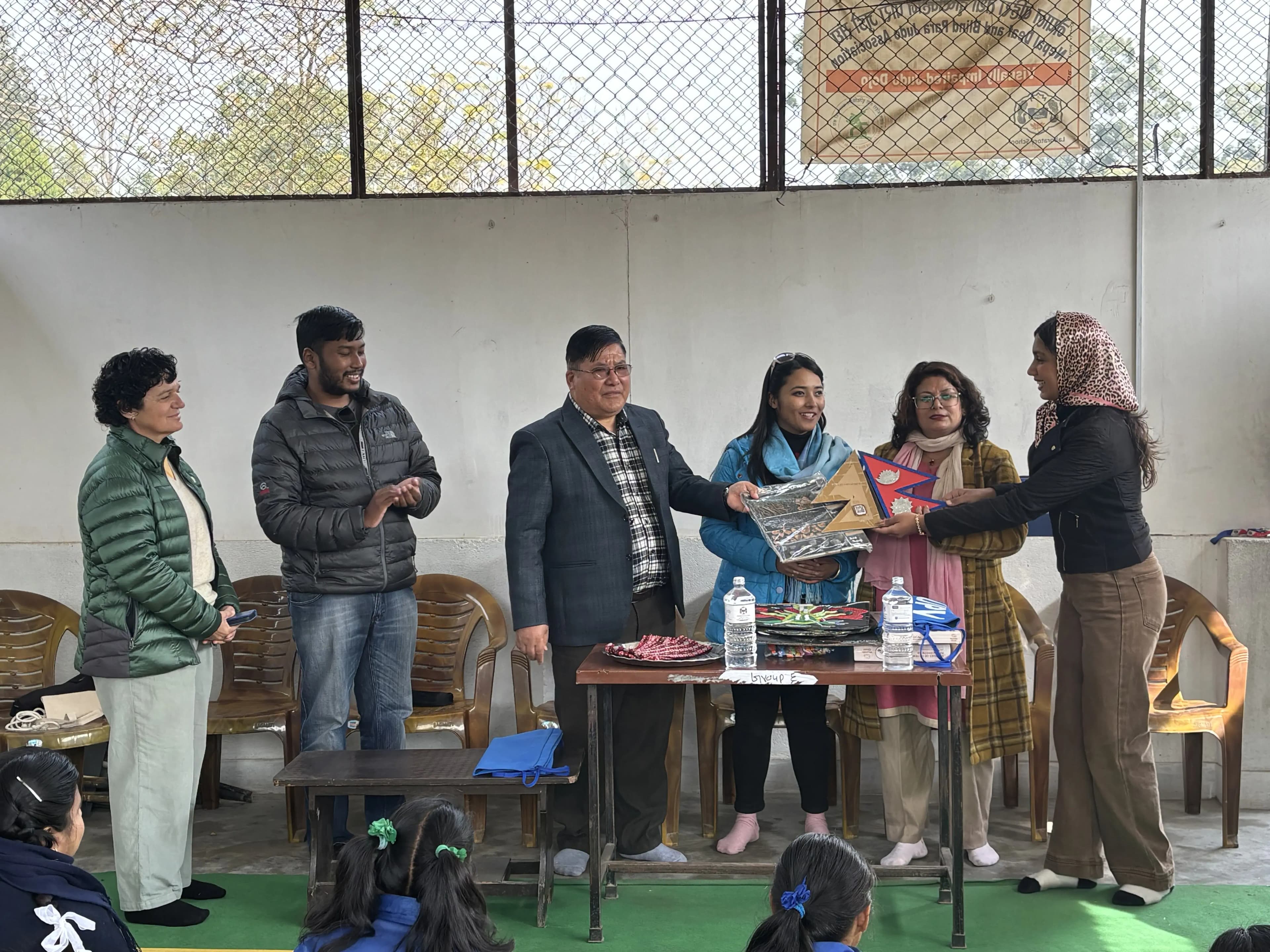 Six adults, including four women and two men, are standing together indoors near a table. The man in the suit and three women are holding a tactile 3D model of the flag of Nepal. They are taking part in a formal event to hand over the accessible 3D model to the representative of the school. The atmosphere is positive, with some participants smiling and others watching attentively.