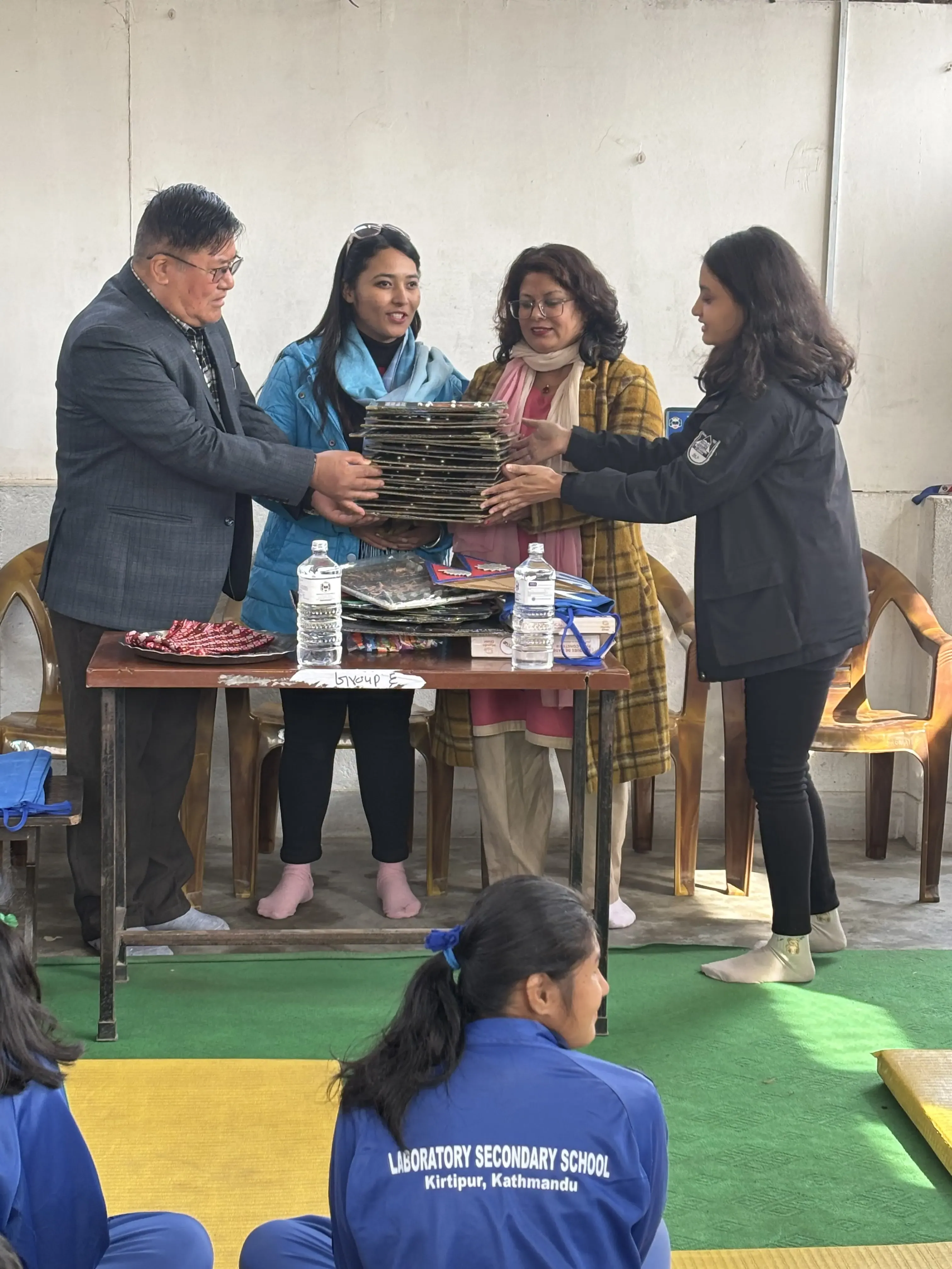 Six adults, including four women and two men, are gathered indoors near a table during a formal event. The man in the suit and three women are holding a stack of tactile 3D models representing the atomic structures of elements. These models are neatly piled one above another. The group is participating in a formal handover ceremony to provide these accessible 3D educational models to the school representatives. In the foreground, students in blue uniforms from Laboratory Secondary School, Kirtipur, Kathmandu, are seated and observing the event.