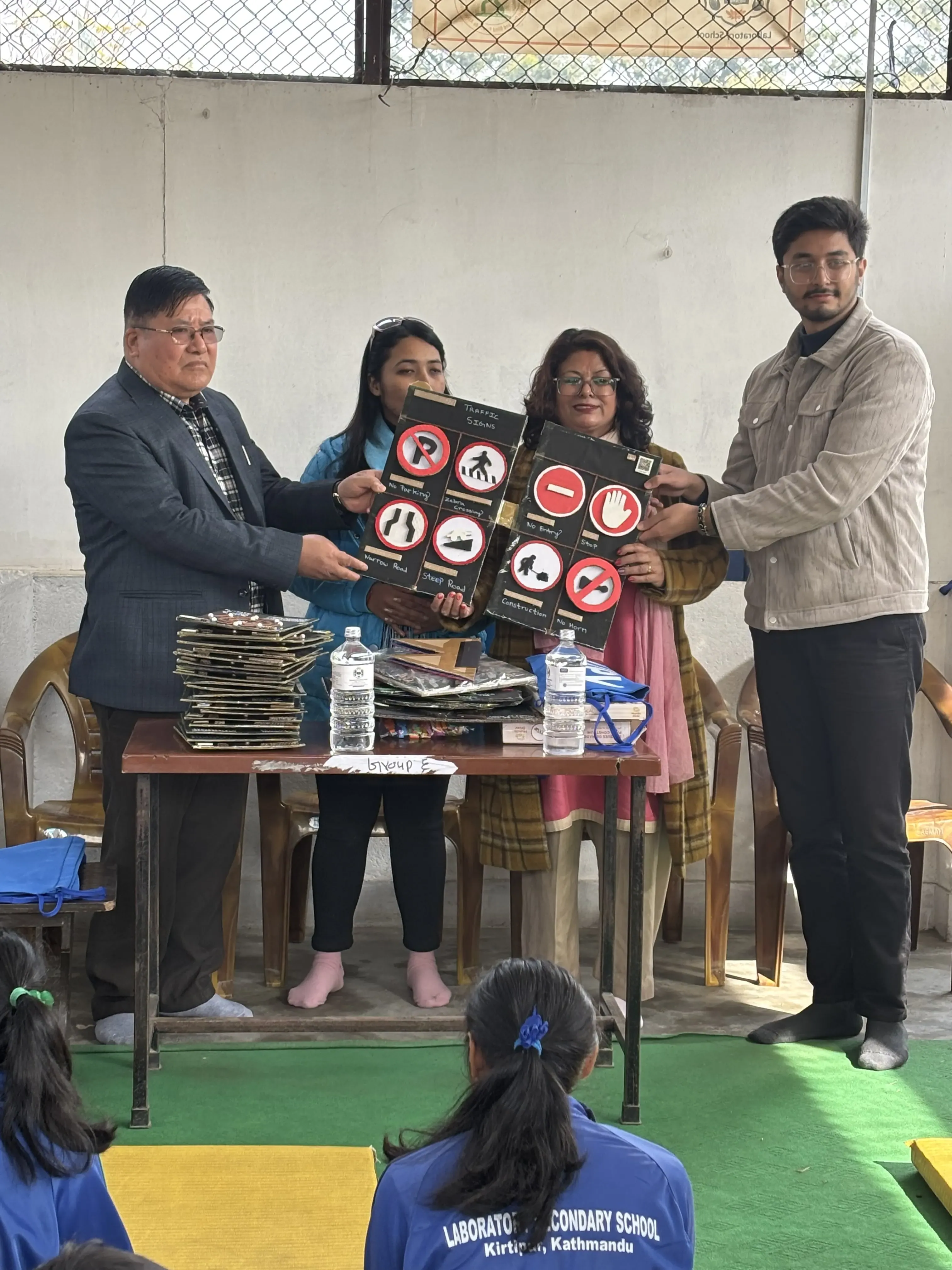 A group of four adults—two men and two women—are standing and sitting near a table indoors. Three of them are holding up large tactile 3D boards displaying traffic signs. On the table in front of them, there are more tactile boards stacked together, along with some bottled water and other items. Two students in blue uniforms from Laboratory Secondary School, Kirtipur, Kathmandu, are seated on the floor nearby, watching the handover event attentively.