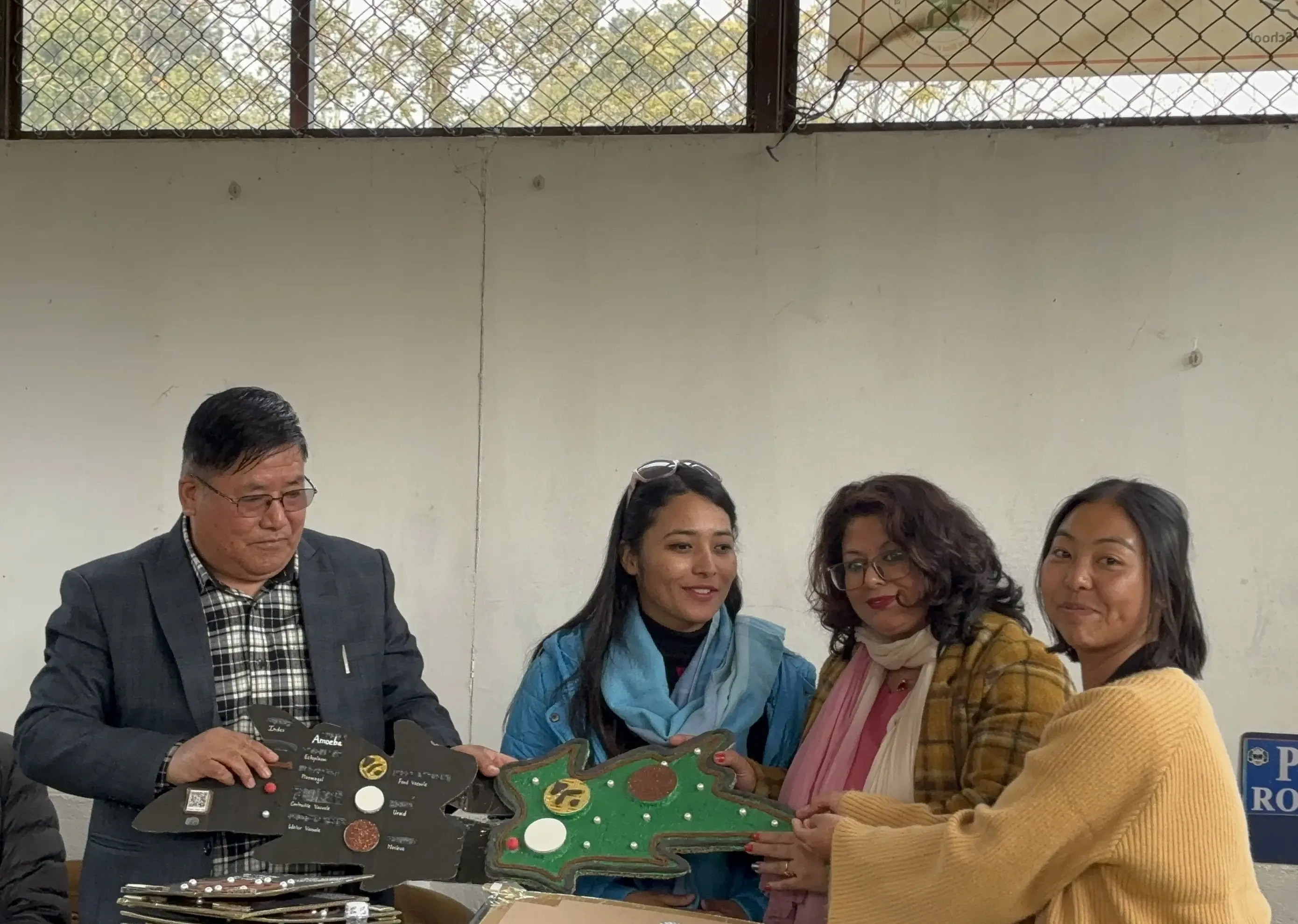 Four adults, one man and three women, are standing together indoors, participating in a formal handover event. The man and one of the women are holding up tactile 3D educational model an amoeba. The women in the center are smiling, and the overall atmosphere is positive and engaged.
